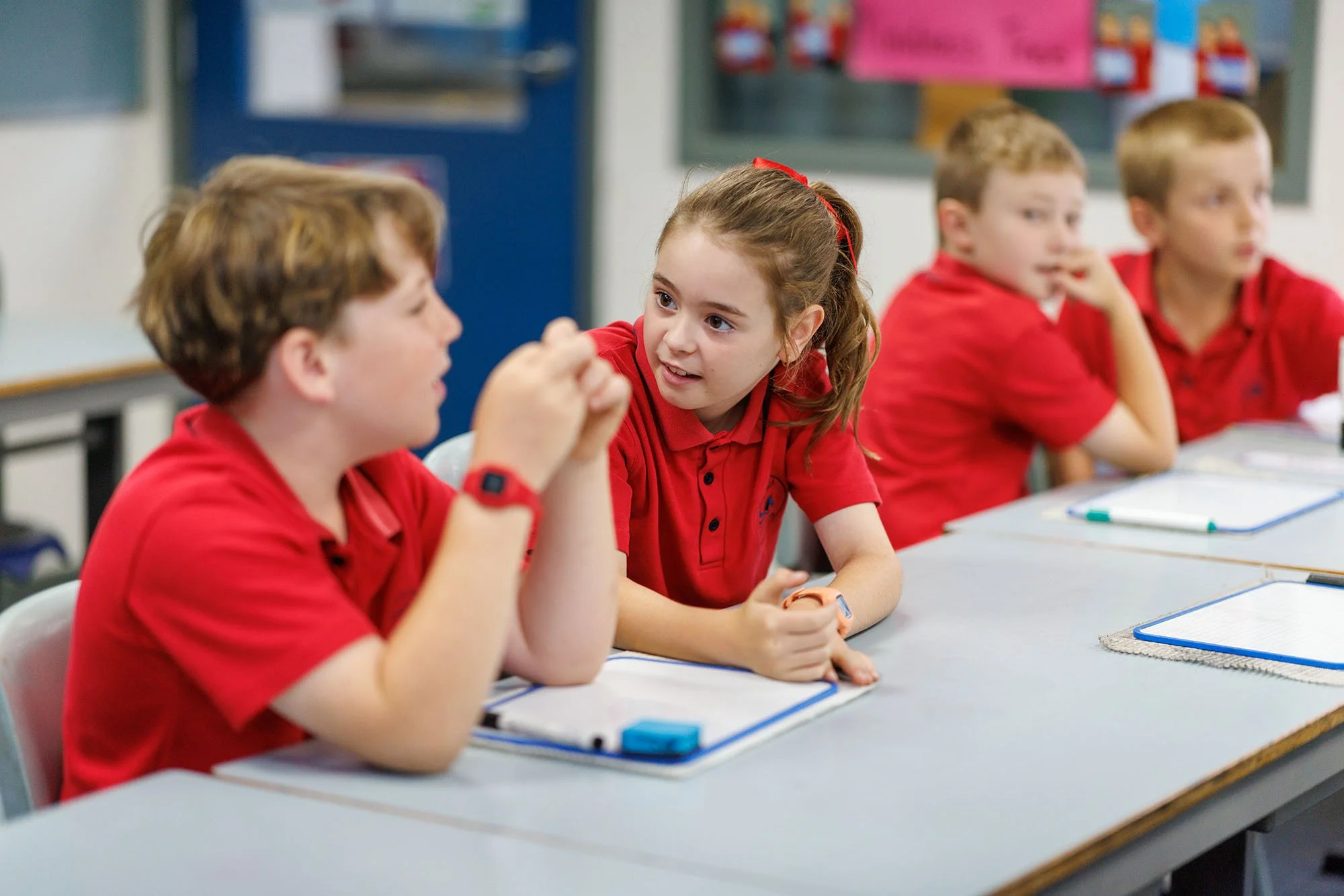 A classroom scene with four students wearing red shirts sitting at desks. The girl in the center is talking to the boy on her left, who is listening and has a wristwatch. The other two students are in the background, one resting his chin on his hand.