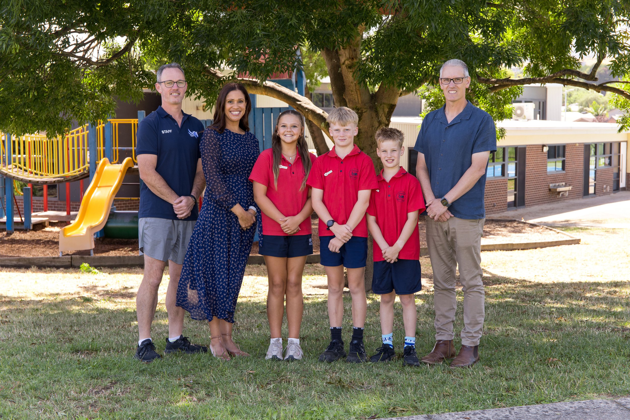 Group of six people, including three adults and three children, standing outdoors under a large tree with a playground in the background. The children are wearing red shirts, and the adults are dressed casually. The scene appears to be at a school or community center.