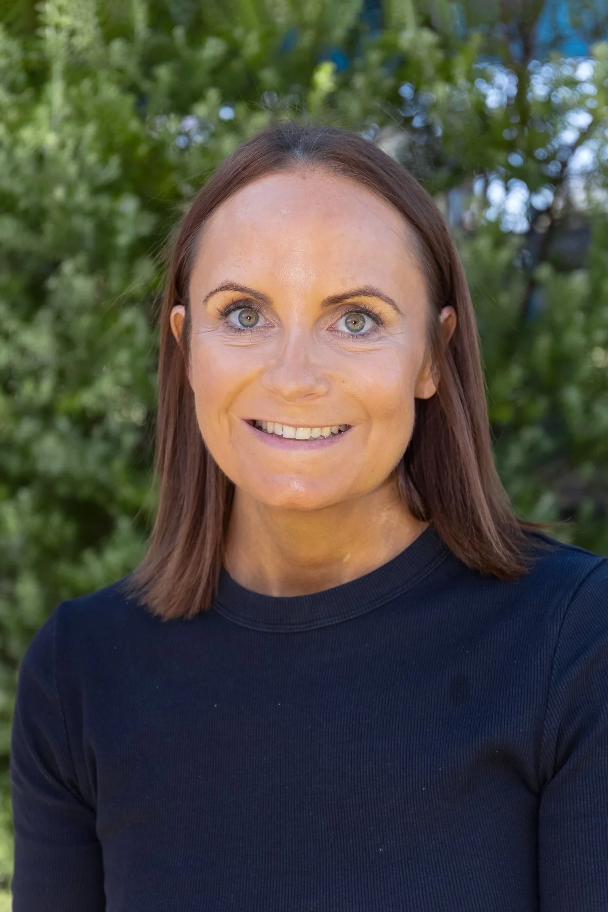 Close-up of a woman with shoulder-length brown hair, blue eyes, wearing a dark blue top, smiling outdoors with green trees in the background.
