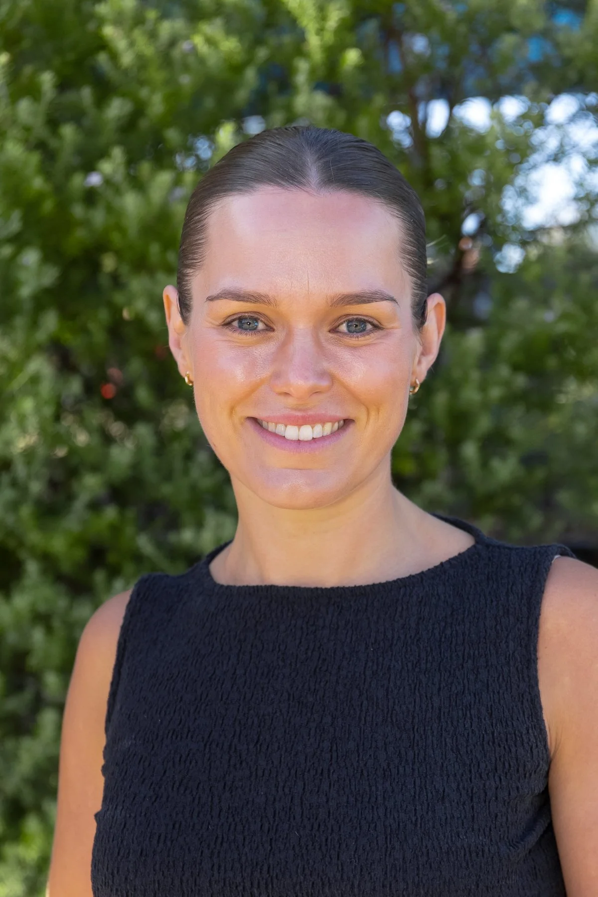A smiling woman with dark brown hair pulled back, wearing a sleeveless dark blue top, standing outdoors with green trees in the background.