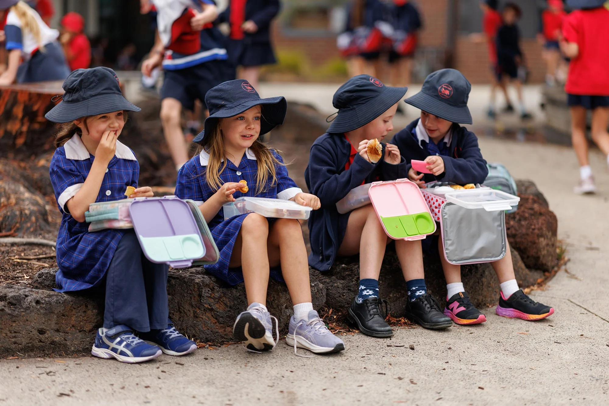 Four school children in uniform sitting on a rock at school yard, eating snacks from lunch boxes, with other children playing in the background.