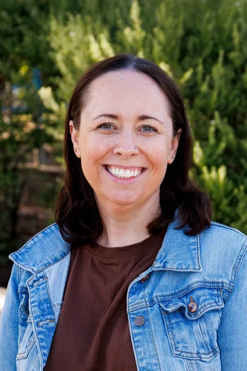 A woman with shoulder-length brown hair smiling outdoors, wearing a denim jacket over a brown shirt, with green trees in the background.