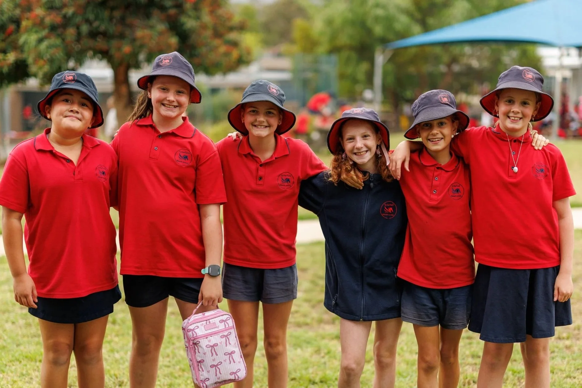 Group of young boys and girls in sports uniforms standing on a blue outdoor sports court, smiling, with some wearing hats and school apparel, likely team members posing for a team photo.