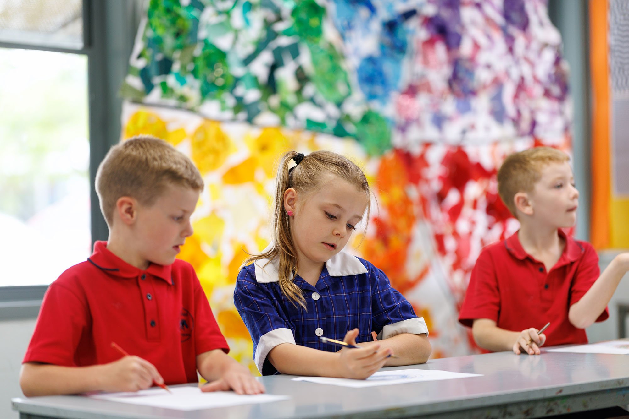 Three children sitting at a table in a classroom. The girl in the middle is writing, and the boys on either side are looking at their papers. The background features colorful, abstract artwork.