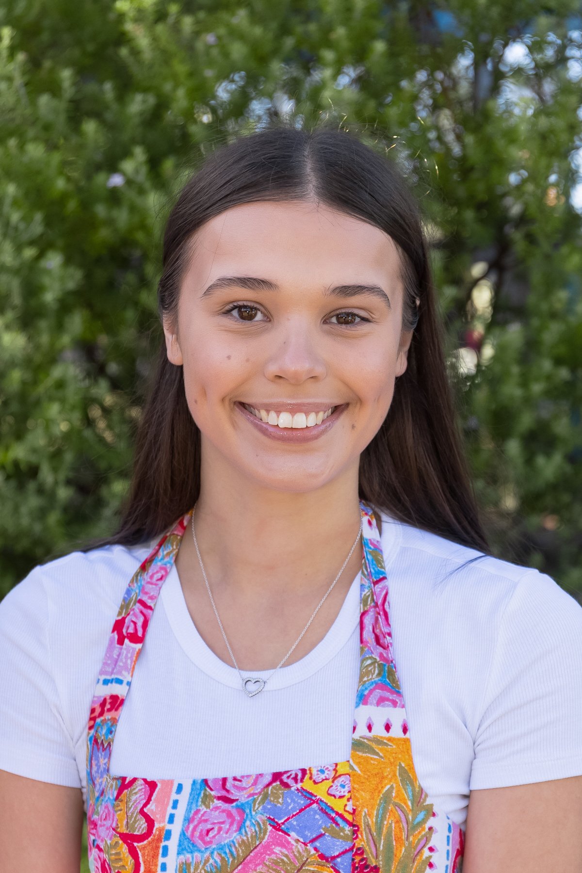 Smiling young woman with long brown hair wearing an apron with colorful floral patterns over a white t-shirt, outdoors with green trees in the background.