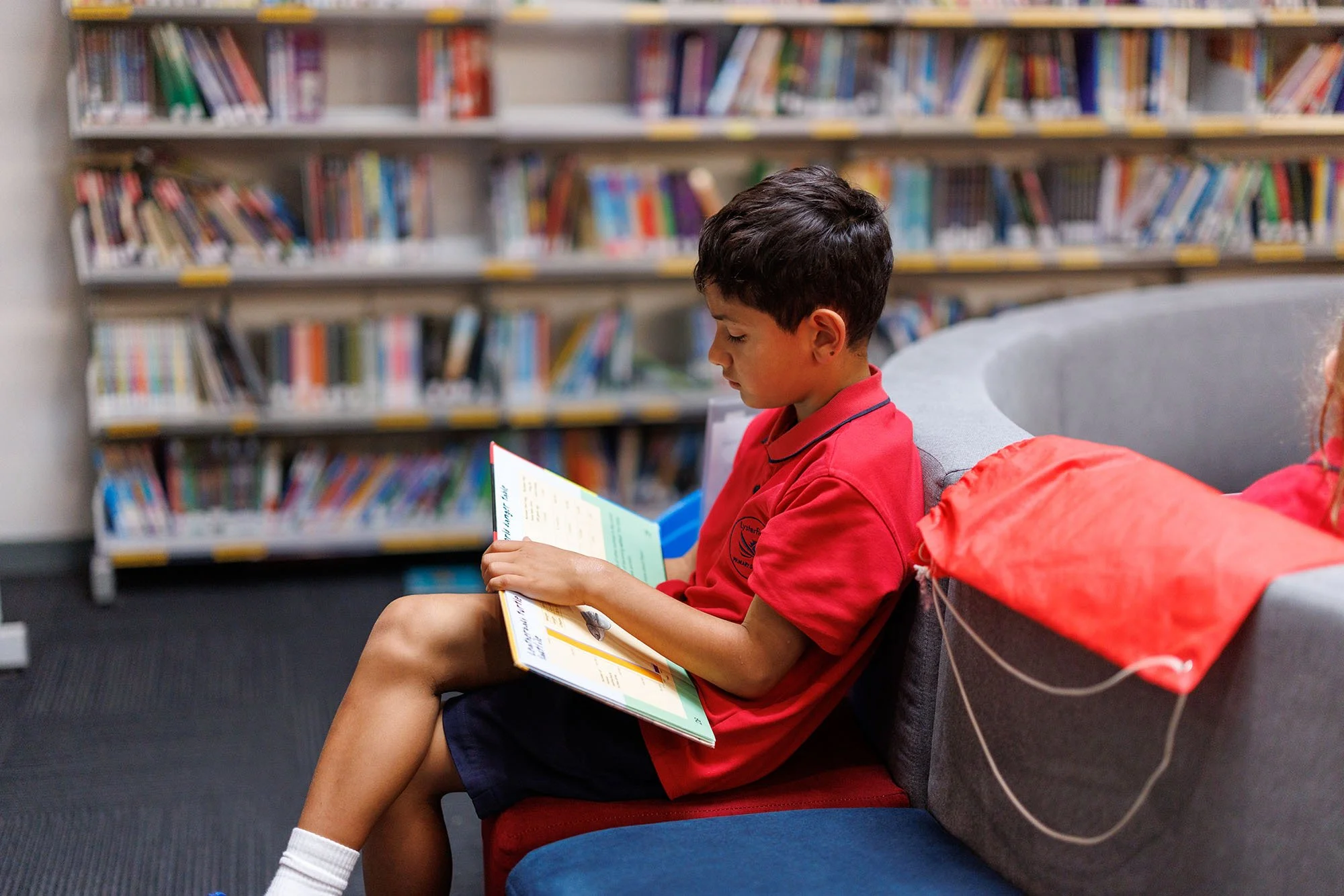 A young boy in a red shirt and dark shorts sitting on a red and grey couch in a library, reading a colorful book.