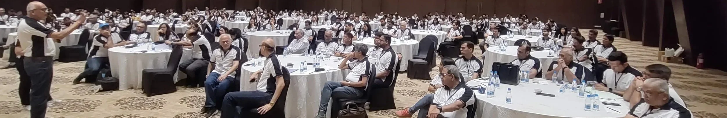 A speaker on stage addressing a large audience seated at round tables in a conference room.