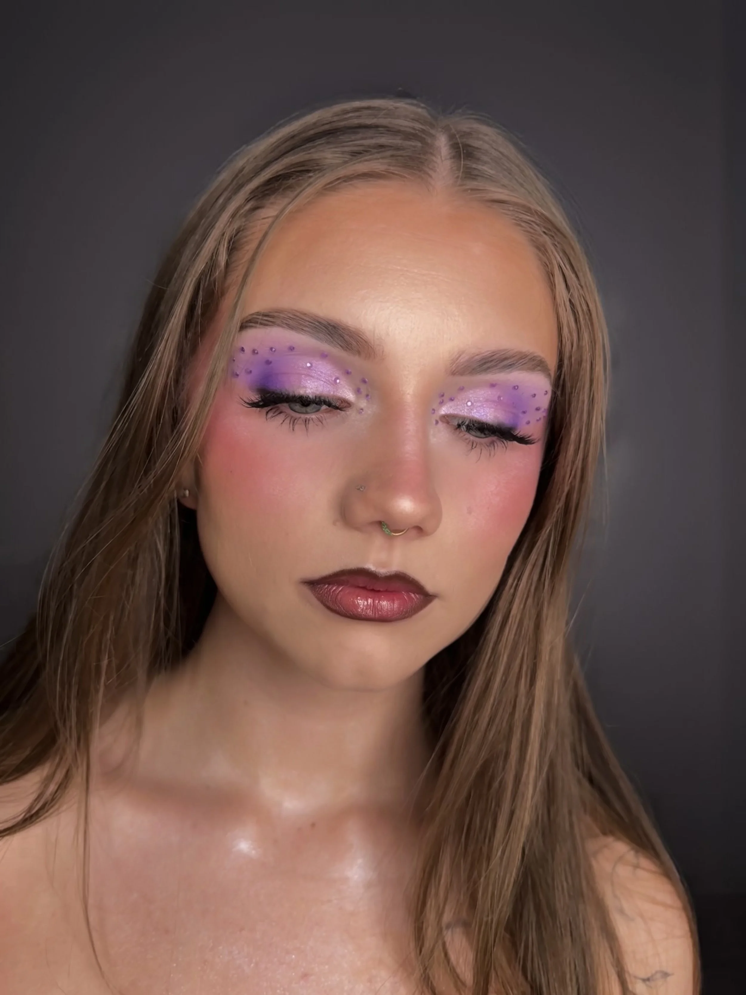 A young woman with long, light brown hair and light skin has closed eyes with purple eyeshadow and small purple rhinestones on her eyelids. She has a septum piercing and is against a dark gray background.