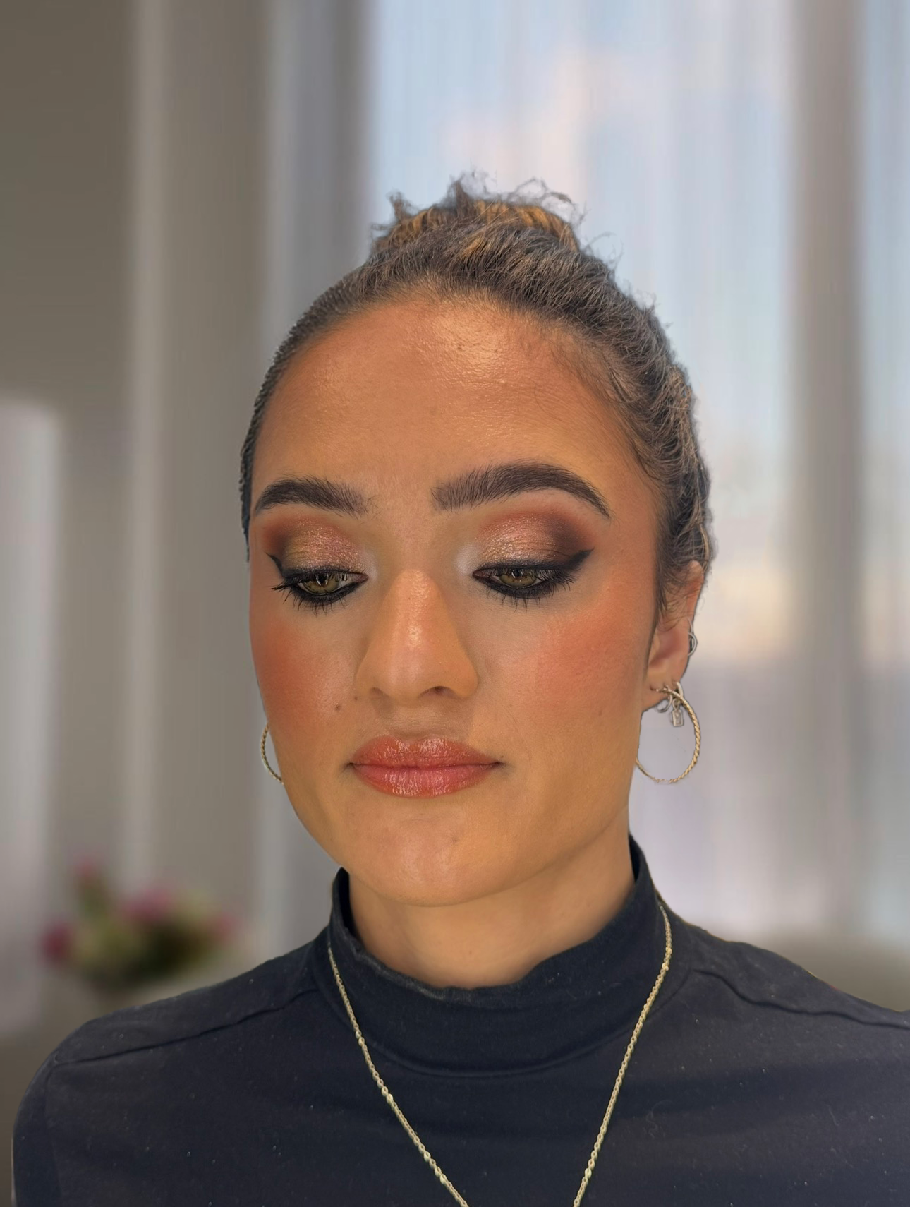 A woman with her hair tied up in a bun, wearing gold hoop earrings, a necklace, and a black turtleneck with makeup including eyeshadow and lipstick.