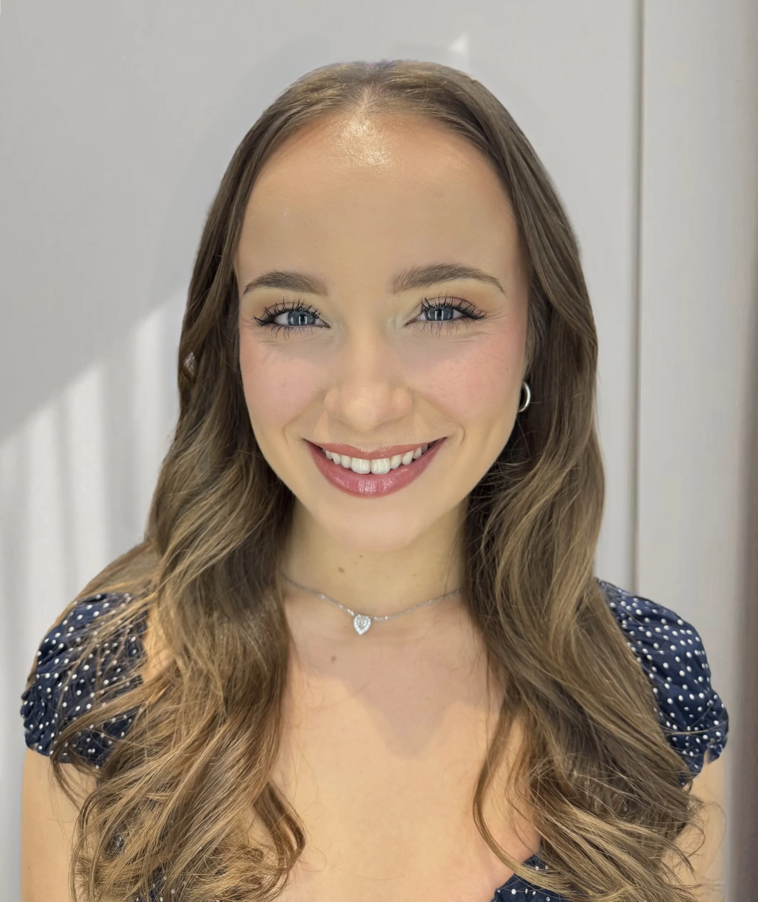 Close-up of a smiling woman with long, wavy brown hair, blue eyes, wearing silver hoop earrings and a heart-shaped pendant necklace, dressed in a navy polka dot blouse with puff sleeves, standing in front of a plain background.