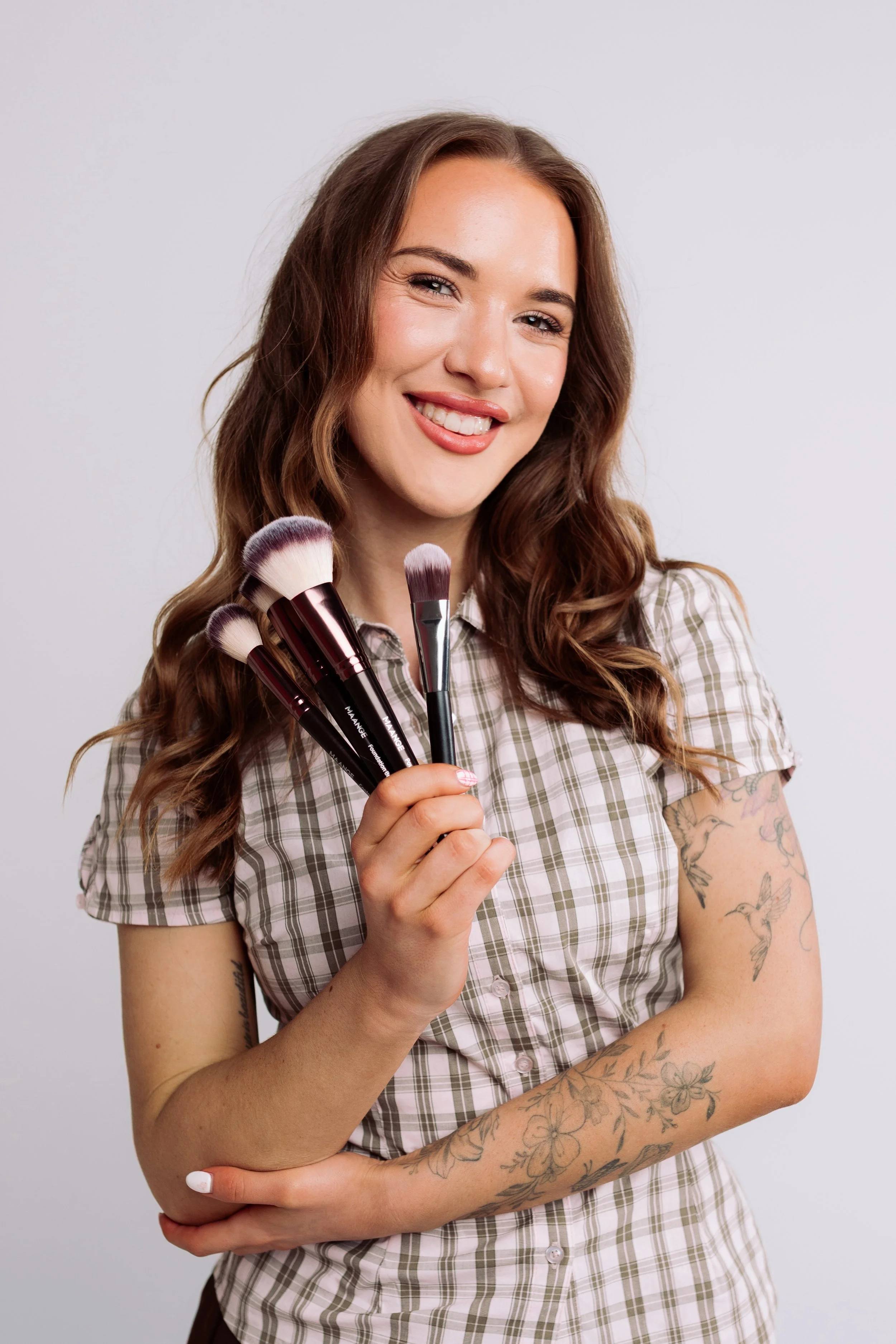 A woman with long wavy brown hair, tattoos on her arms, and a plaid shirt, smiling and holding several makeup brushes in front of a plain light gray background.