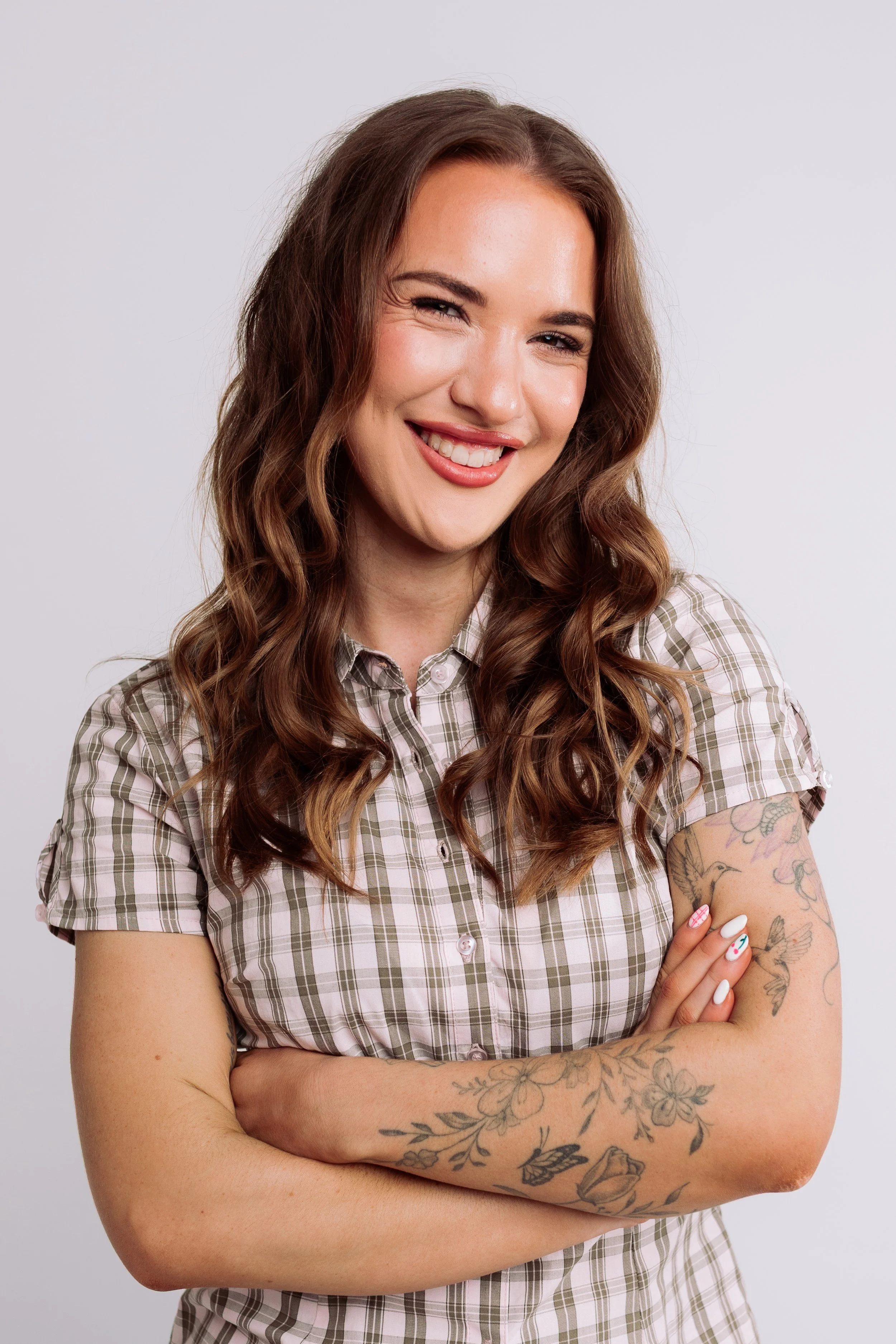 A woman with long, wavy brown hair, smiling with a closed-mouth smile, wearing a plaid short-sleeve shirt, arms crossed, and tattoos on her right arm, standing against a plain white background.