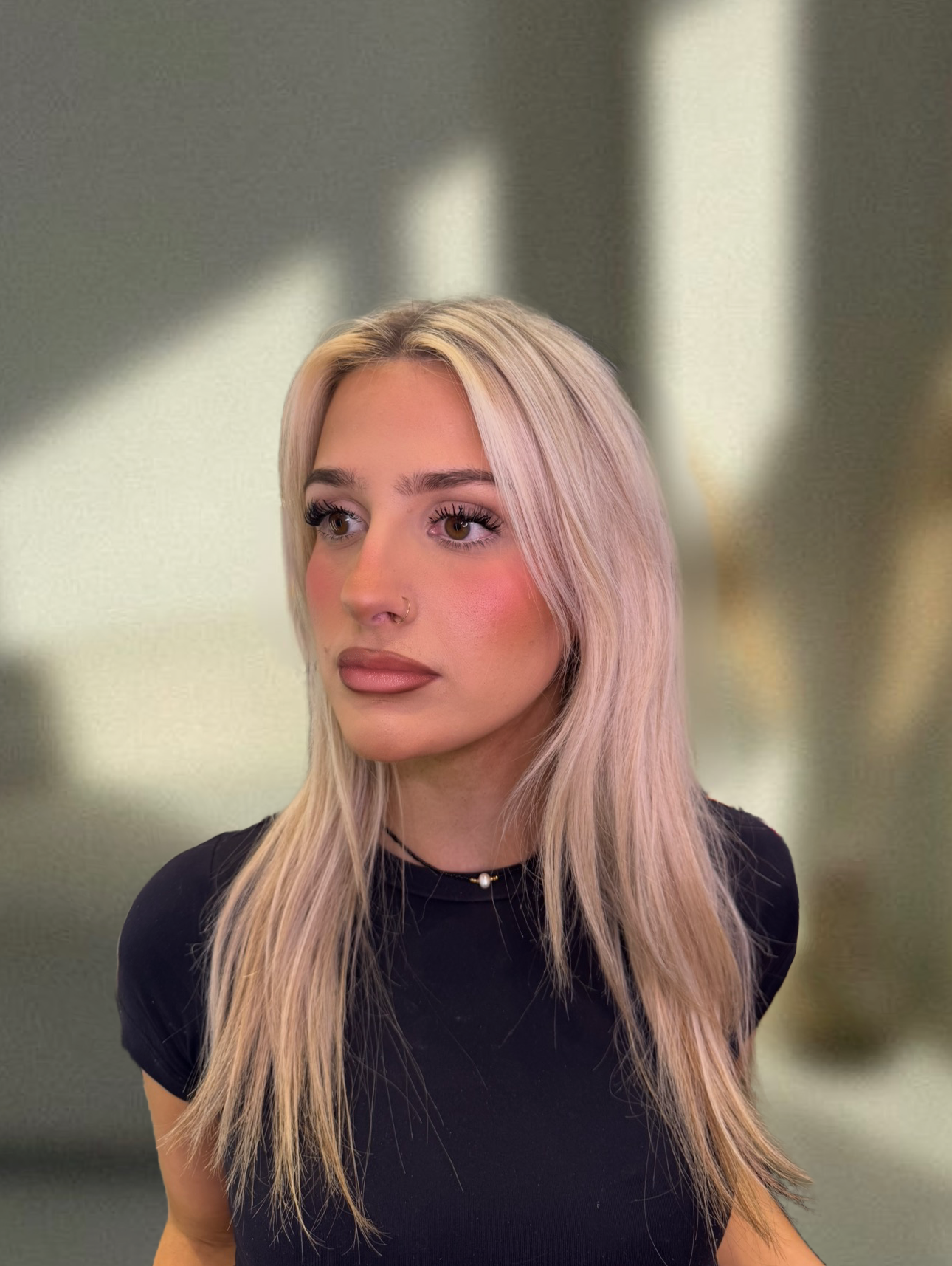 A young woman with long blonde hair, wearing a black top and a choker necklace with a pearl, looks to her right against a blurred background with vertical light and dark stripes.