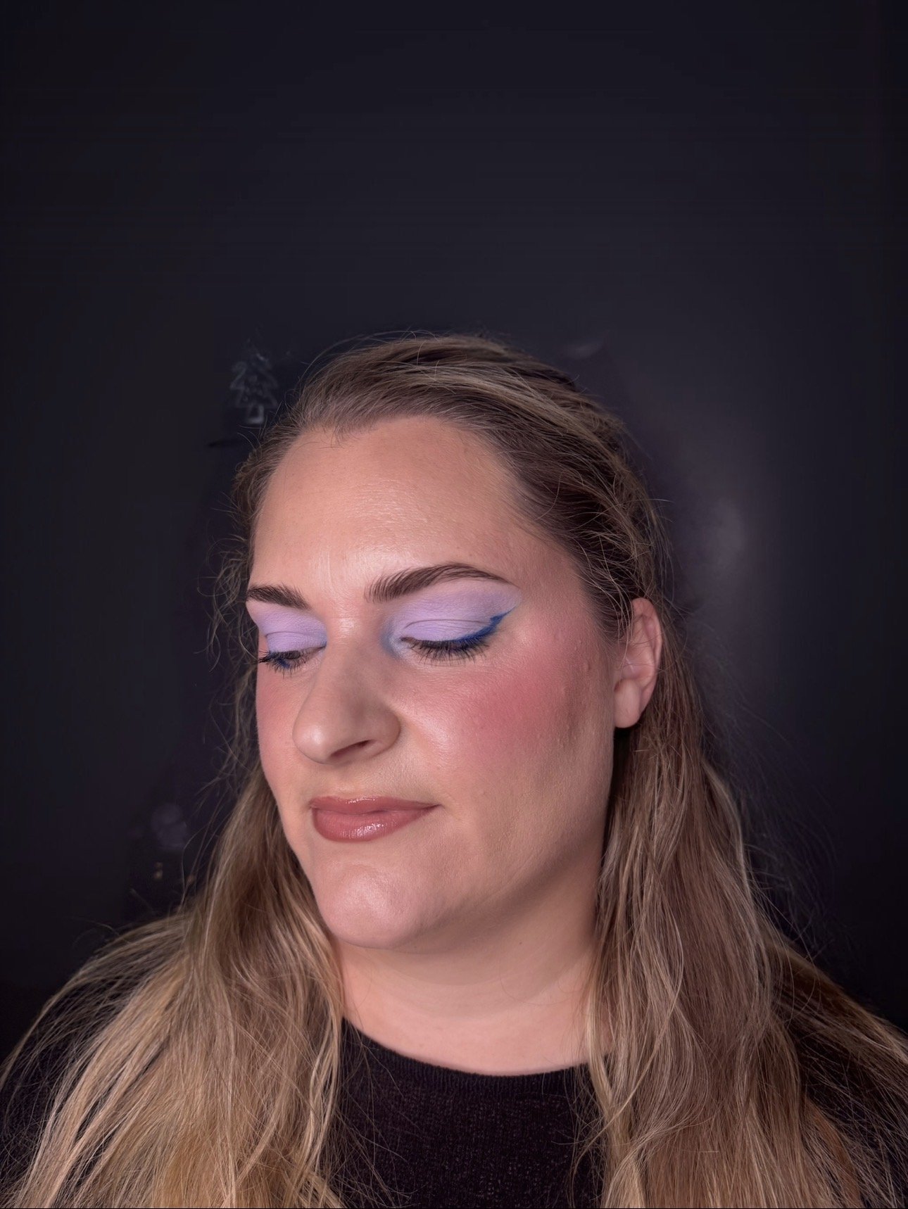 A woman with light skin and long wavy hair is shown with her eyes closed, showcasing her colorful eye makeup that includes shades of purple and blue eyeshadow with blue eyeliner, and a natural lips makeup. The background is solid black.