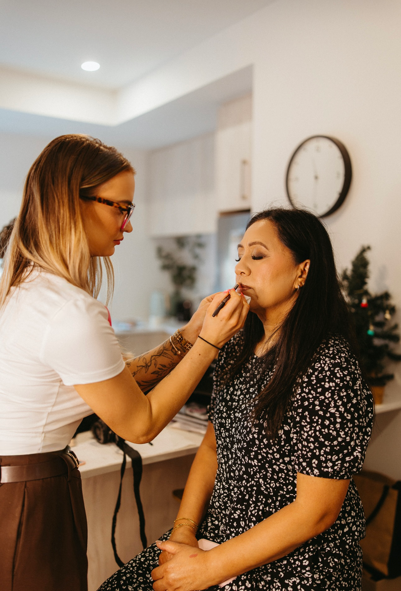 A woman sitting with her eyes closed while another woman applies makeup to her lips in a well-lit room decorated for the holidays, with a Christmas tree in the background.