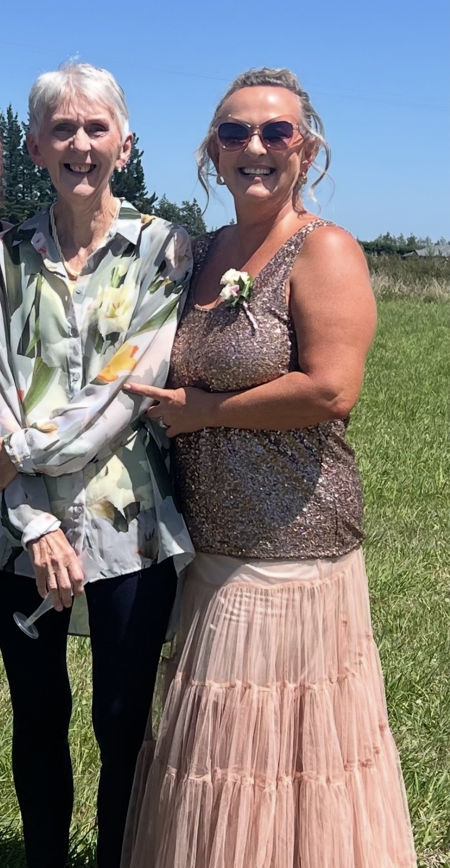 Two women smiling outdoors on a sunny day, dressed in semi-formal attire, with greenery and clear blue sky in the background.
