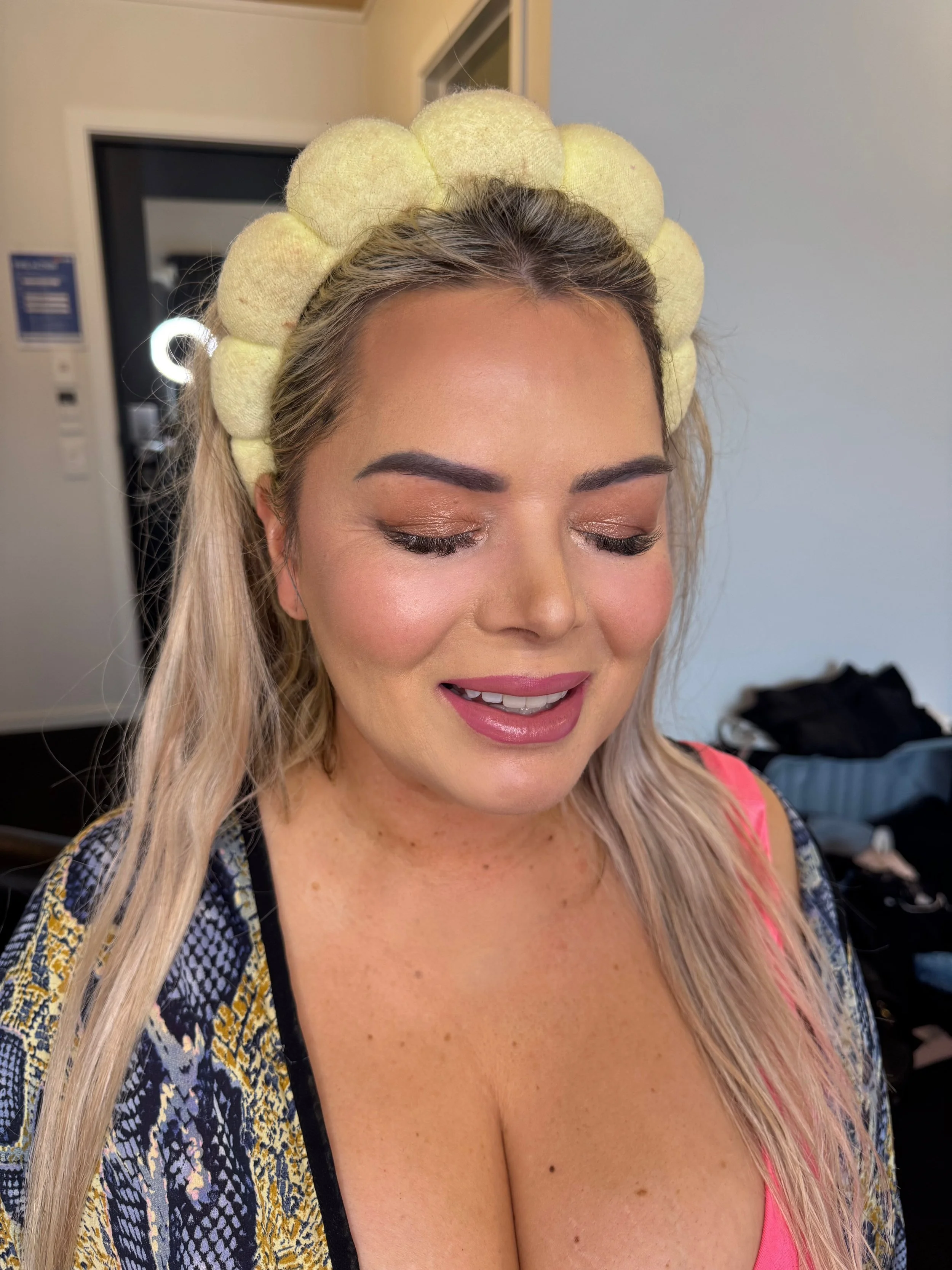 A woman with blonde hair and light skin smiling with her eyes closed, wearing a yellow hair net made of rolled fabric puffs and a colorful patterned top. She appears to be in a dressing room or backstage area.