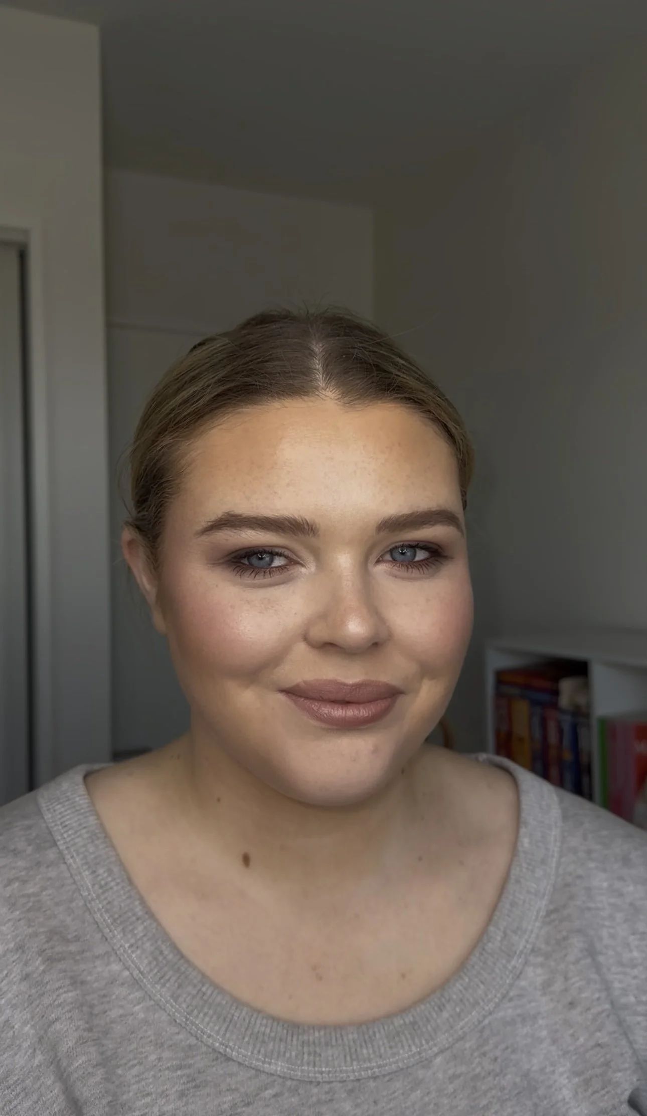 Close-up of a smiling woman with light brown hair, blue eyes, wearing a gray sweater, and a background featuring a white wall, bookshelf with books, and a partially visible closet door.