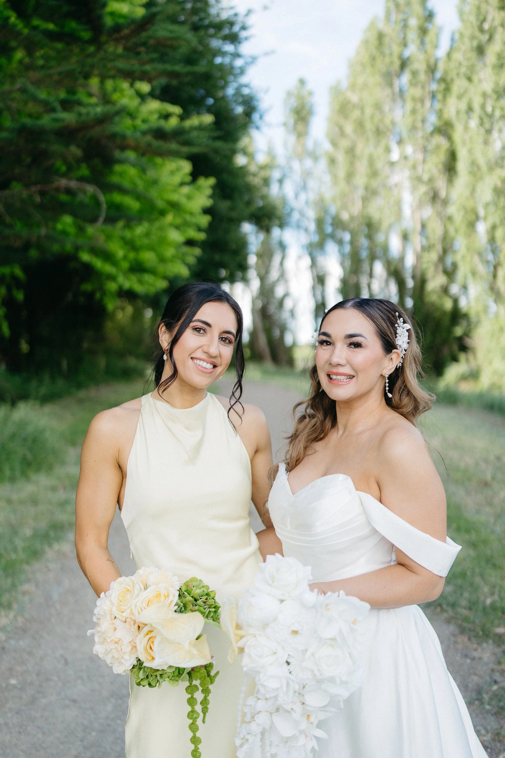 Two women smiling and standing outdoors on a path surrounded by trees, one in a light cream dress and the other in a white wedding gown holding a bouquet of white and cream flowers with green accents.