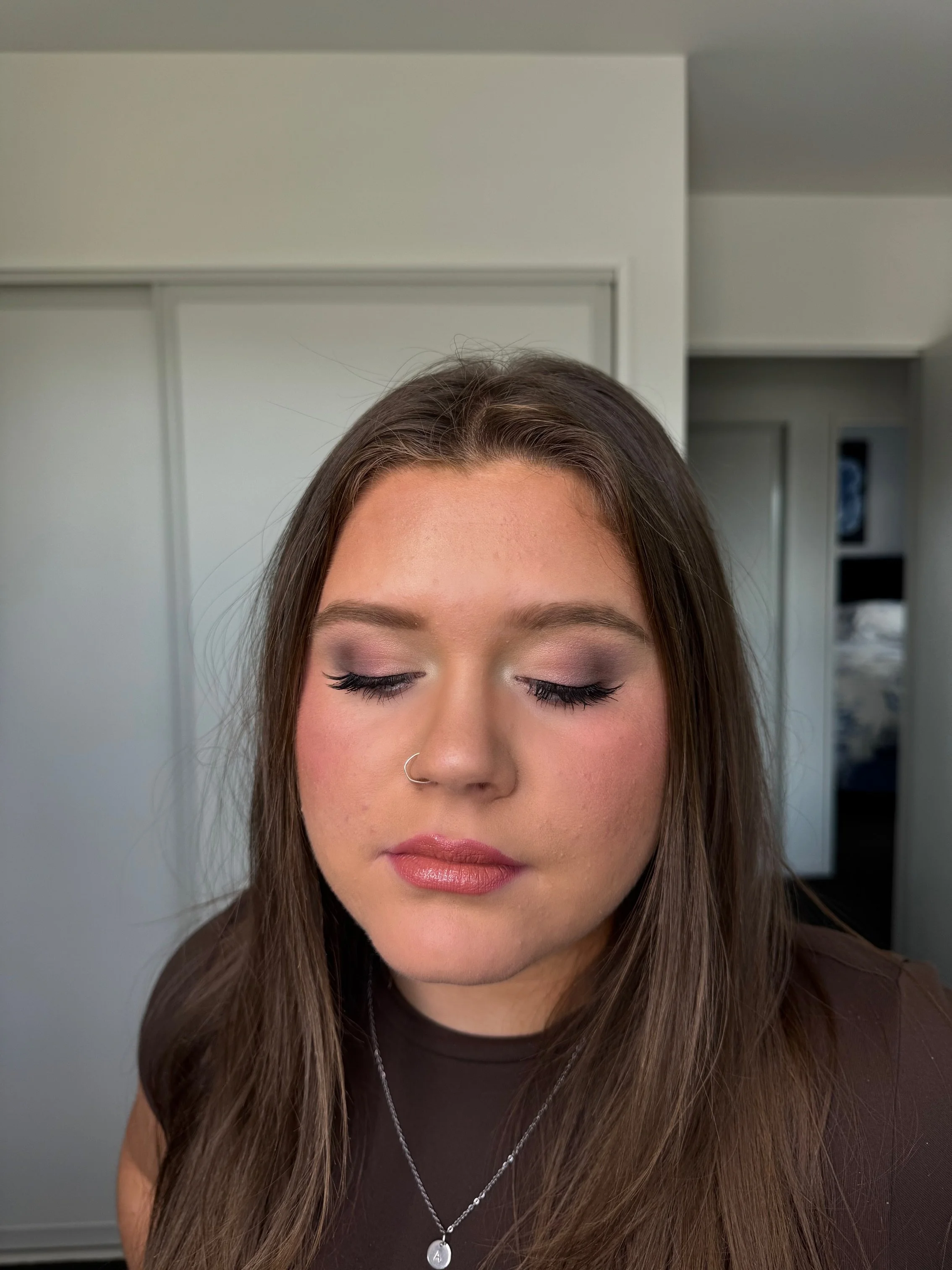 Close-up of a young woman with long brown hair, closed eyes, and makeup, standing in a room with white walls and a door in the background.