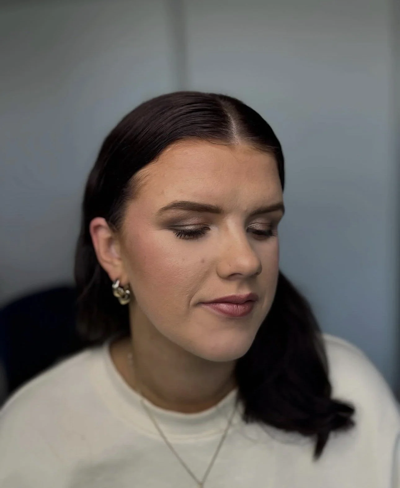 A woman with dark brown hair and fair skin, wearing earrings and a white top, has her eyes closed and appears to be resting or in contemplation against a light blue wall.