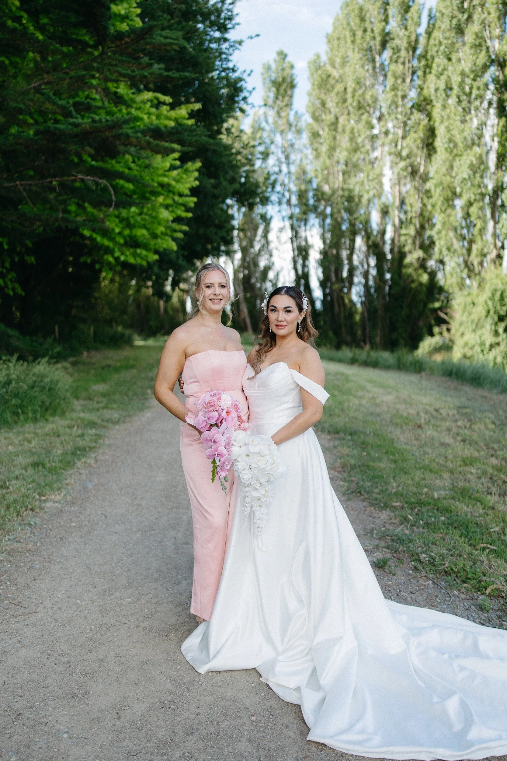 Two women in wedding dresses standing on a dirt path surrounded by green trees, smiling at the camera.