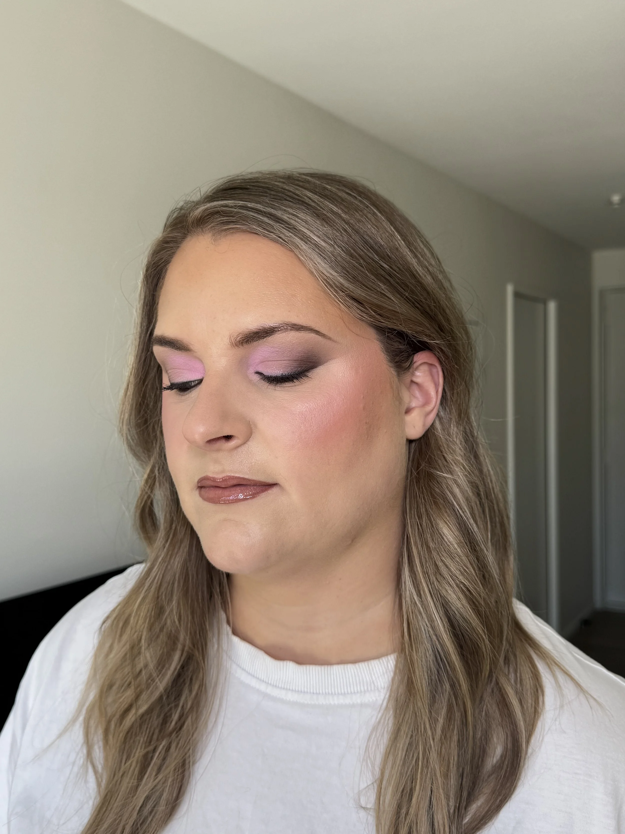 A woman with long, wavy blonde hair and subtle makeup with pink eyeshadow and glossy lips, wearing a white top.