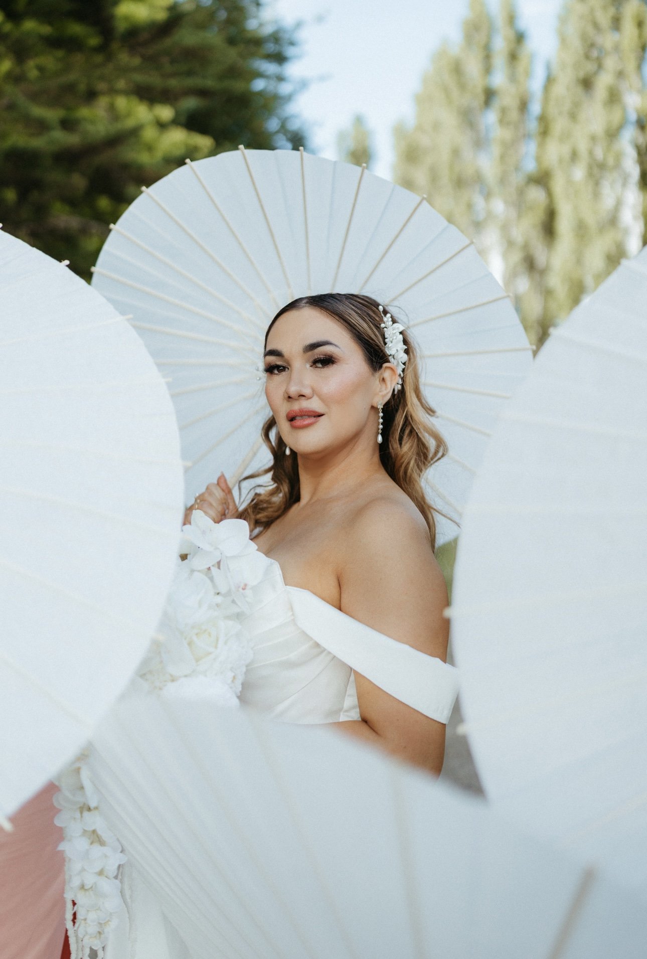 A woman in a white off-the-shoulder wedding dress holding white parasols outdoors, with trees and a blue sky in the background.