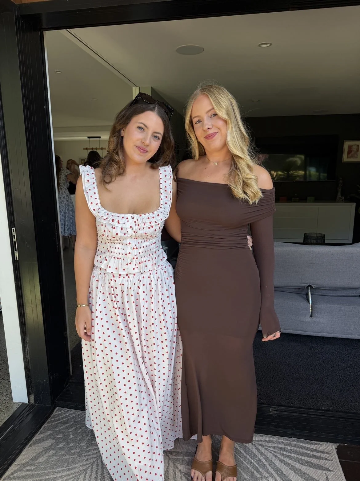 Two women standing side by side in a house, one wearing a white dress with red polka dots and sunglasses on her head, the other wearing a brown off-shoulder dress, both smiling at the camera.
