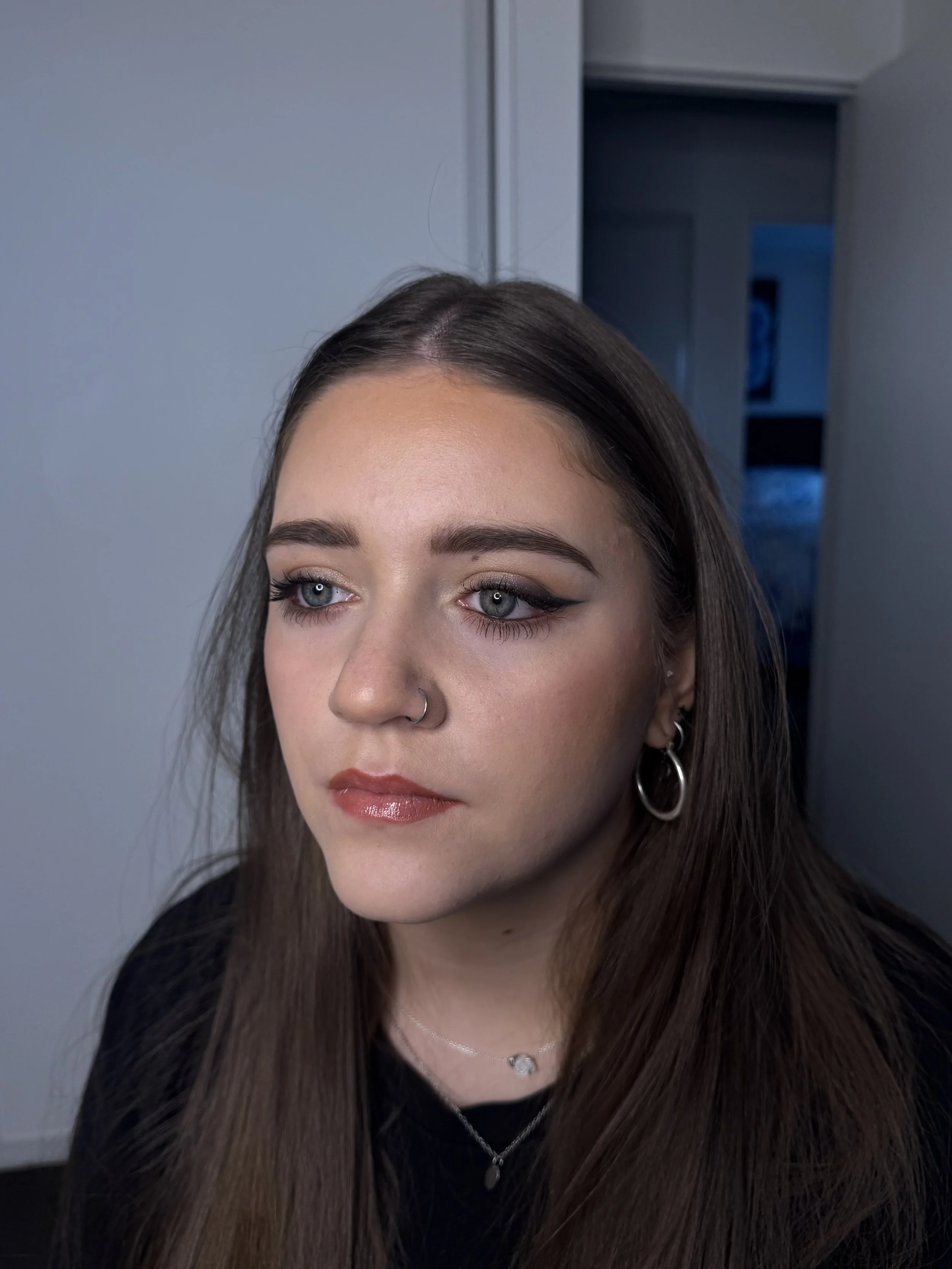 Close-up of a woman with long brown hair, blue eyes, makeup, earrings, and a nose ring, indoors with a light blue wall and doorway in the background.