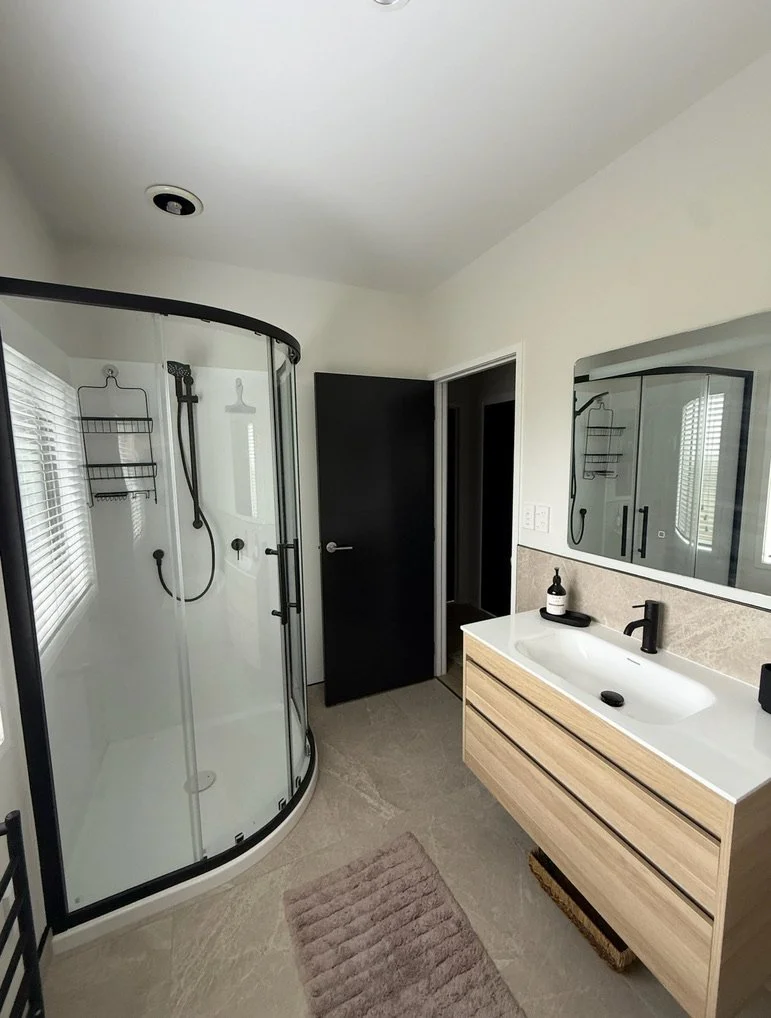 Bathroom with a glass shower enclosure, a white vanity with a black faucet, and a large mirror. The room has beige tiled floors, a pink bath mat, and a window with blinds.