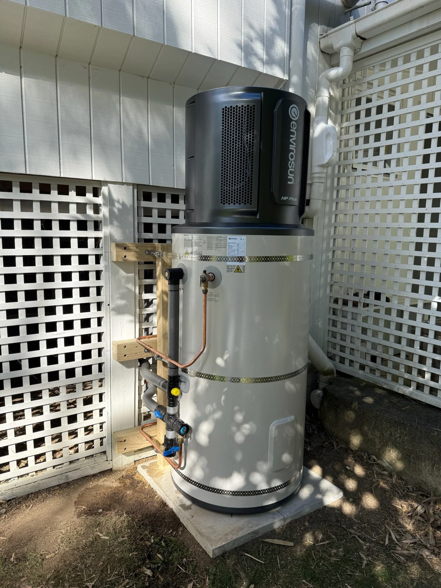 An outdoor tankless water heater system with pipes and equipment mounted on a concrete slab, installed next to a lattice fence and a building wall.