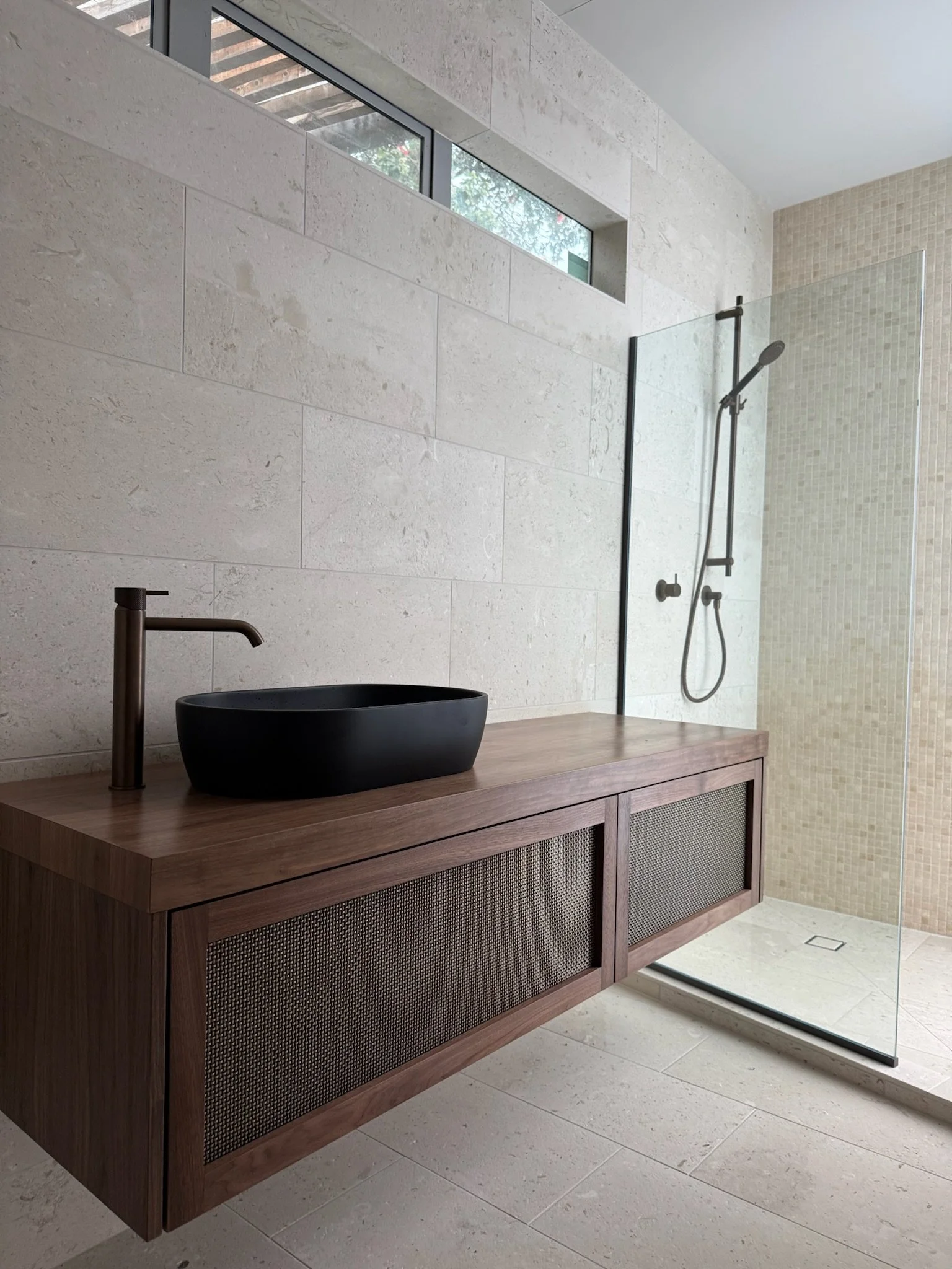 Modern bathroom with a black vessel sink on a wooden vanity, a bronze faucet, and a walk-in shower with a glass door and pebble tile walls.