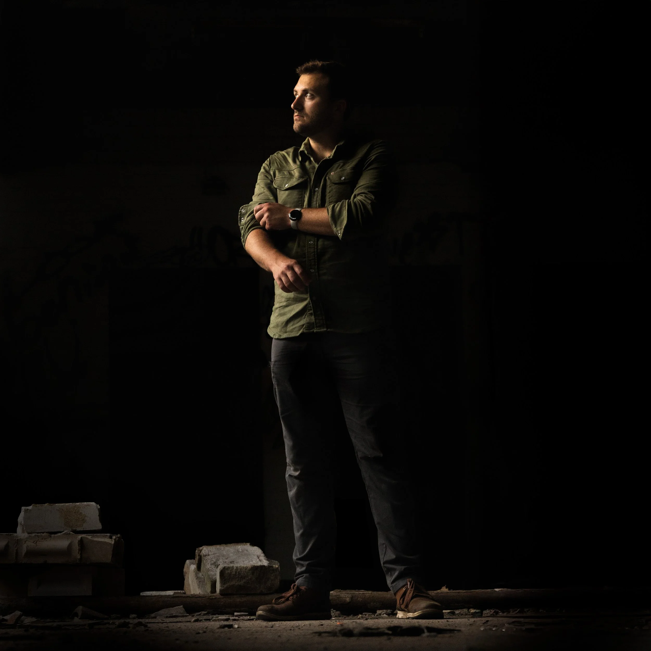 James Stevens standing with cross-armed posture in a dark, abandoned room with bricks on the ground and a faint graffiti in the background.
