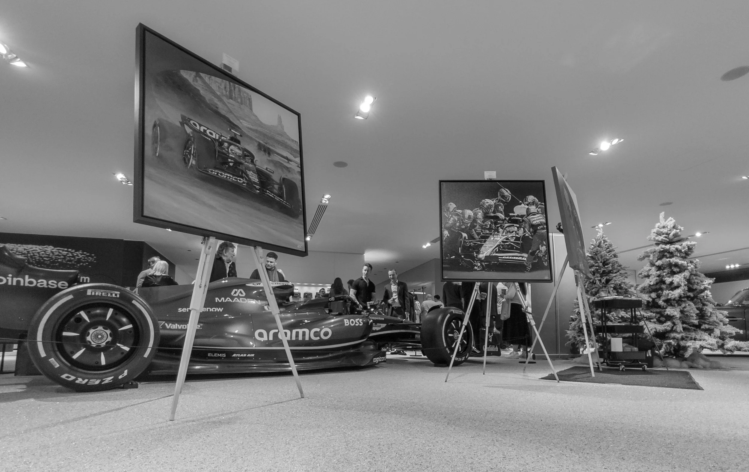 Black and white photo of a Formula 1 Aston Martin race car exhibition with two large displayed photographs of racing cars on easels, people observing, and holiday trees in the background.