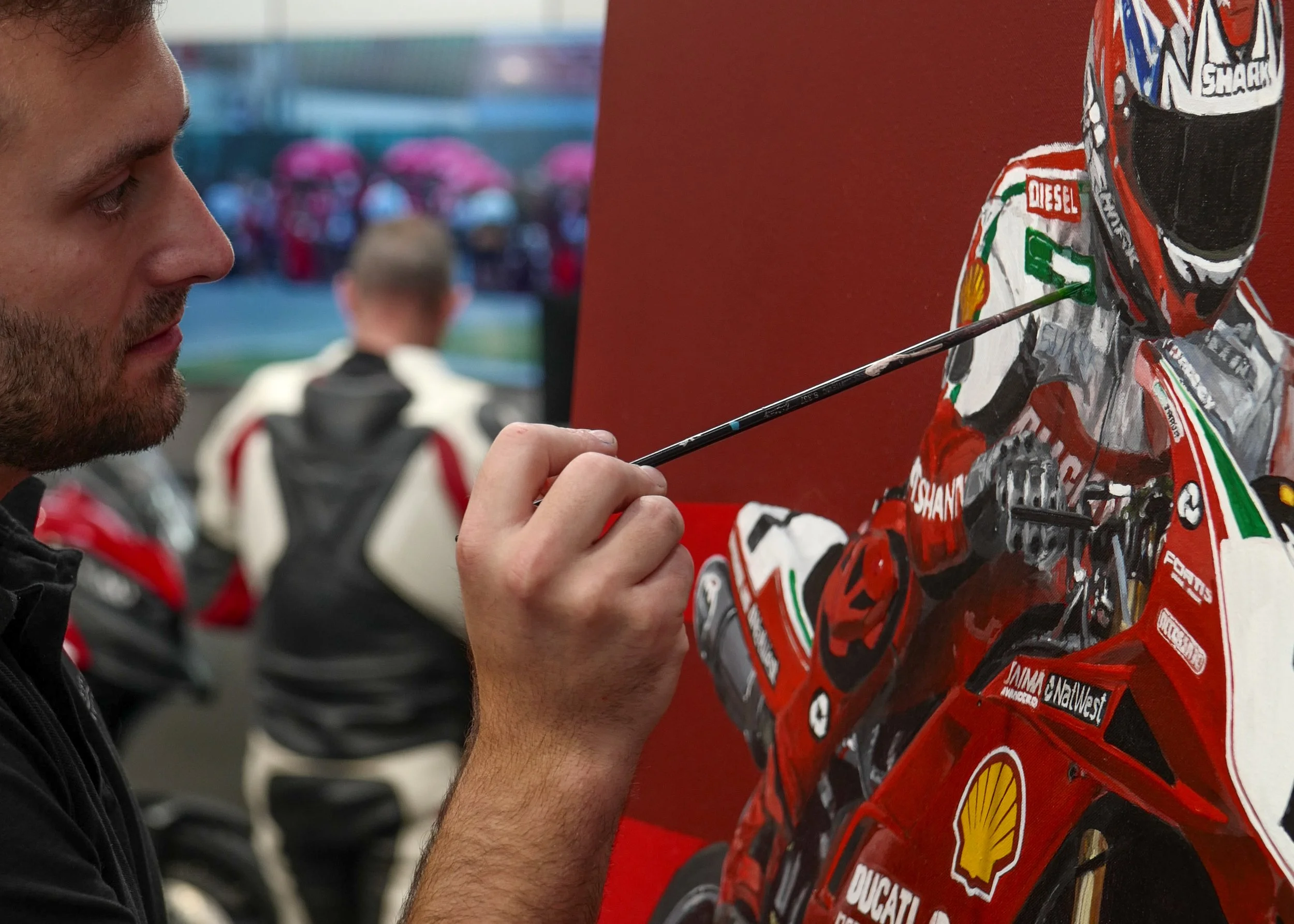 James Stevens painting a detailed motorcycle racing scene on a canvas with Carl Fogarty and Ducati, with motorcycle gear visible in the background.