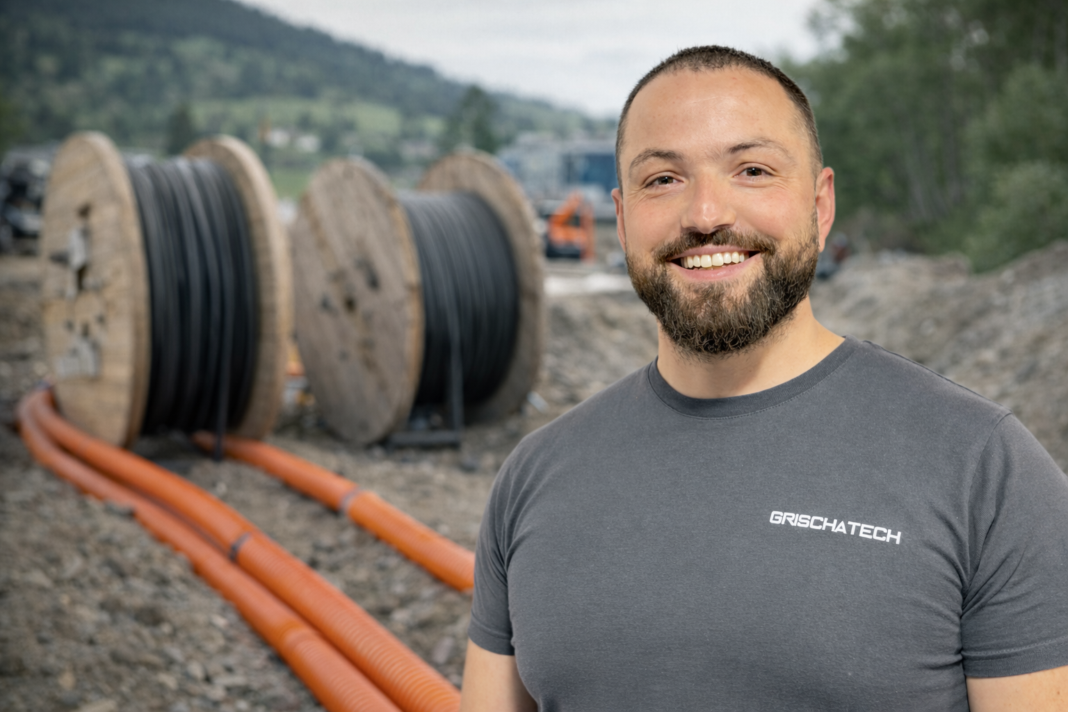 Ein lächelnder Mann steht vor einer Baustelle mit Kabelrollen und orange Leitungen im Hintergrund.