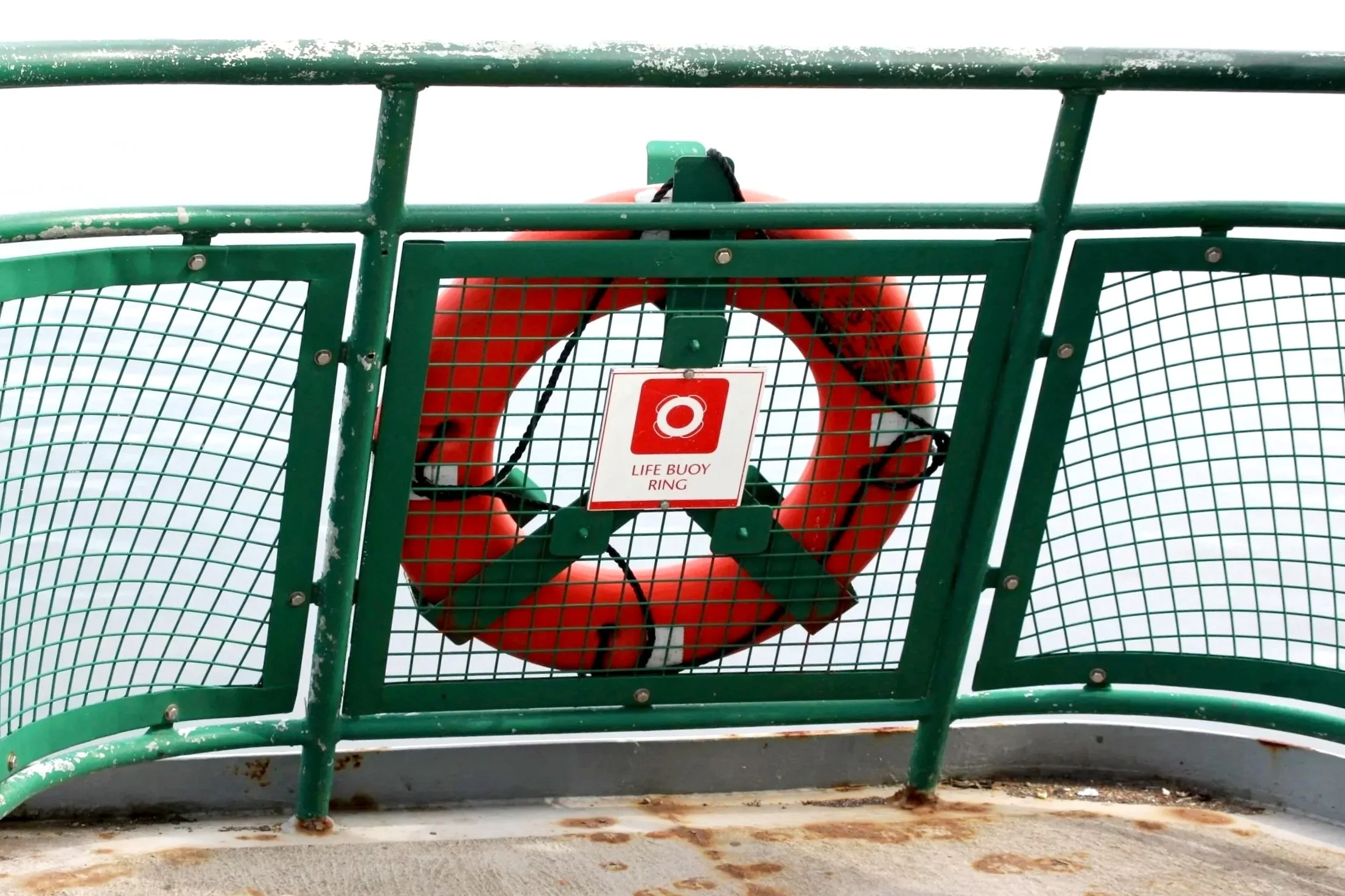 A green safety railing surrounds a circular orange life buoy ring with a white sign that reads 'LIFE BUOY RING' hanging on it, on a boat deck with rust stains.