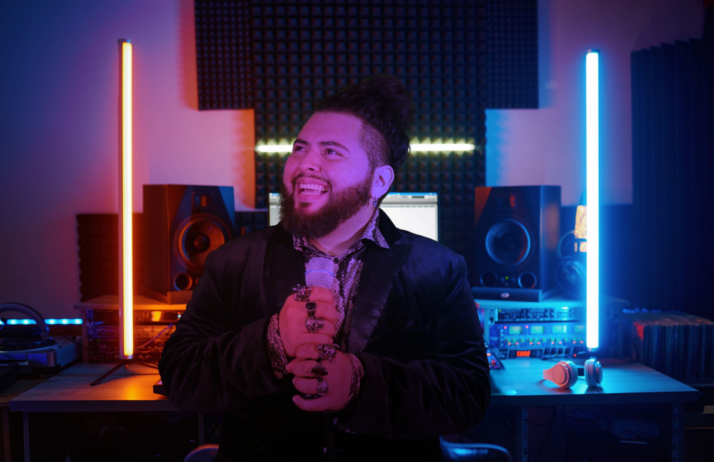 A man with a beard and styled hair, wearing a black blazer and patterned shirt, holding a microphone in a recording studio illuminated with multicolored LED lights. The studio has speakers, audio equipment, and soundproofing foam.