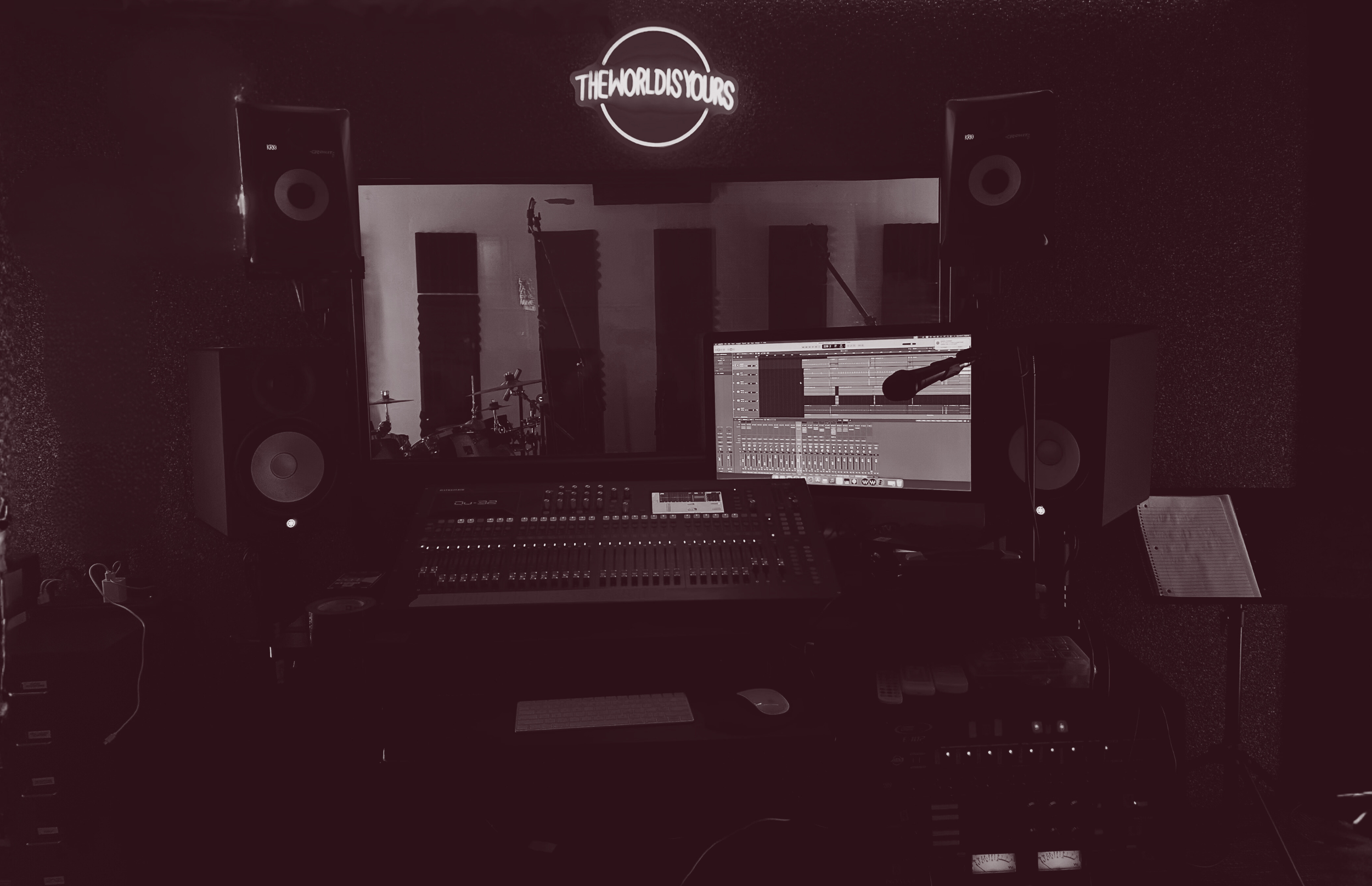 A music recording studio with large speakers, a computer screen displaying audio tracks, a mixing console, and a sign that reads 'The World is Yours' on the wall.