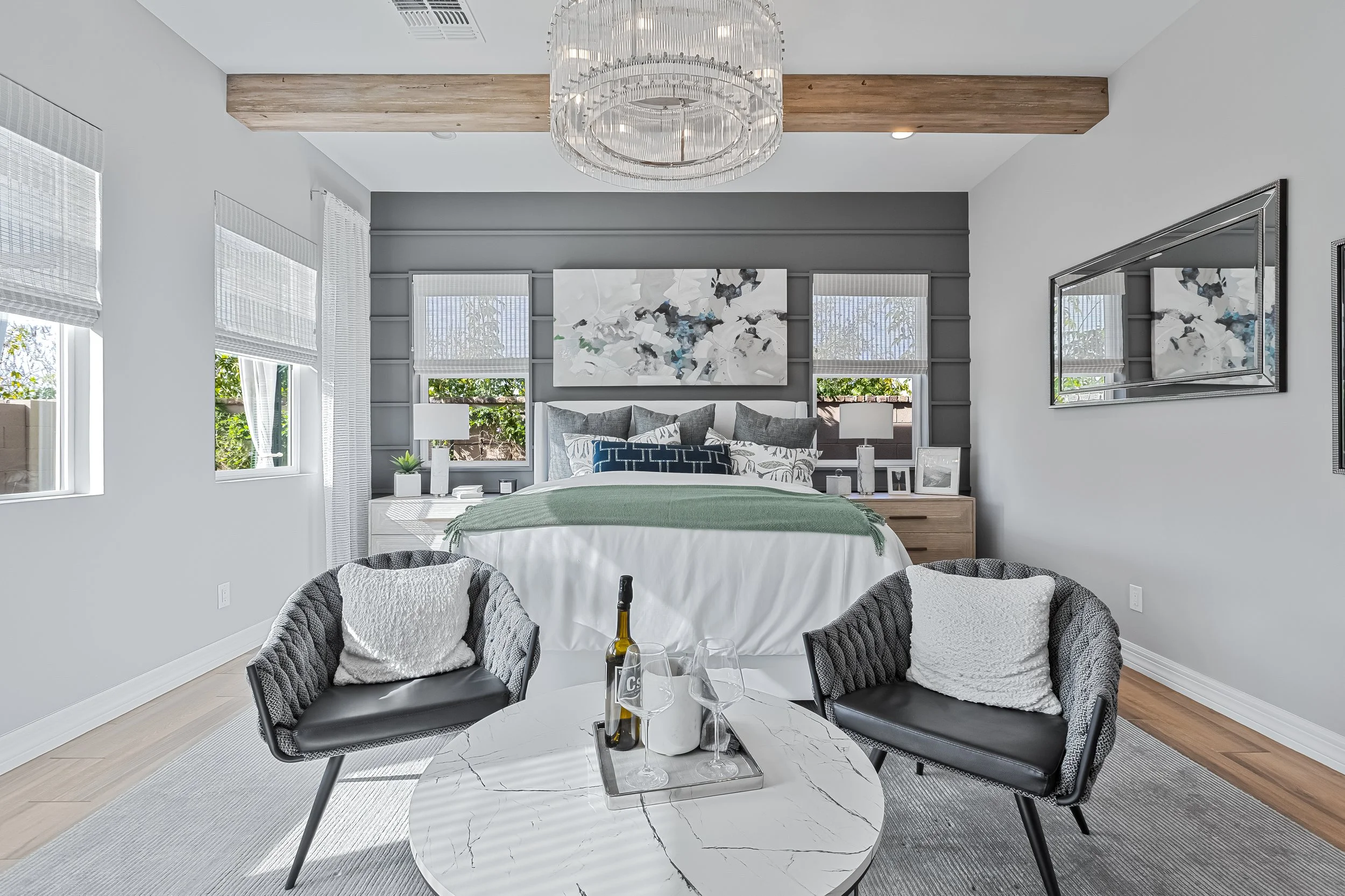 Modern bedroom with a gray and white color scheme, featuring a bed with multiple pillows, two windows behind, a nightstand with decorative items, and a seating arrangement with two black chairs and a round marble table with wine bottles and glasses.