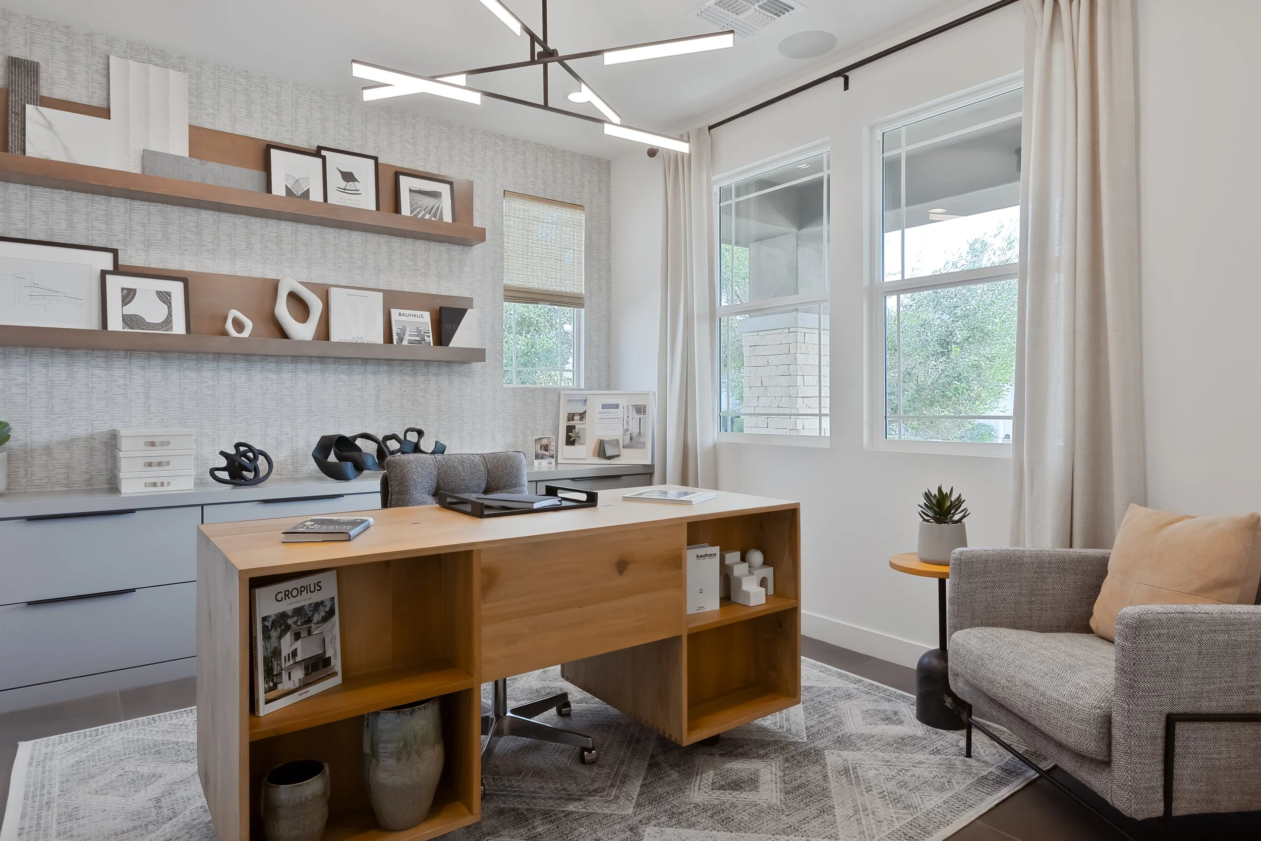 Modern home office with a wooden desk, gray chair, gray couch with beige pillow, and a small side table with a potted plant. White walls, large windows with curtains, and artistic decor on built-in shelves.
