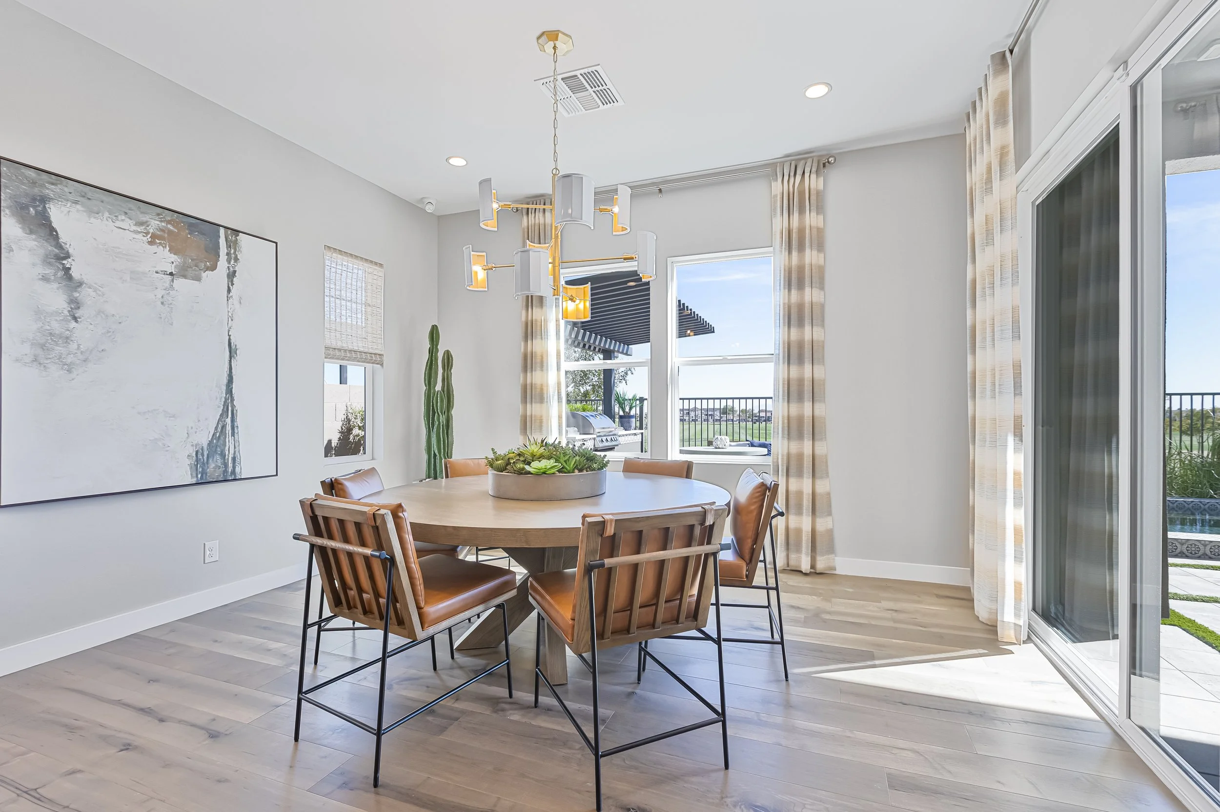 Modern dining room with a wooden round table, six leather chairs, large abstract artwork on the wall, a tall cactus plant, large windows with beige curtains, and a view of an outdoor patio and blue sky.