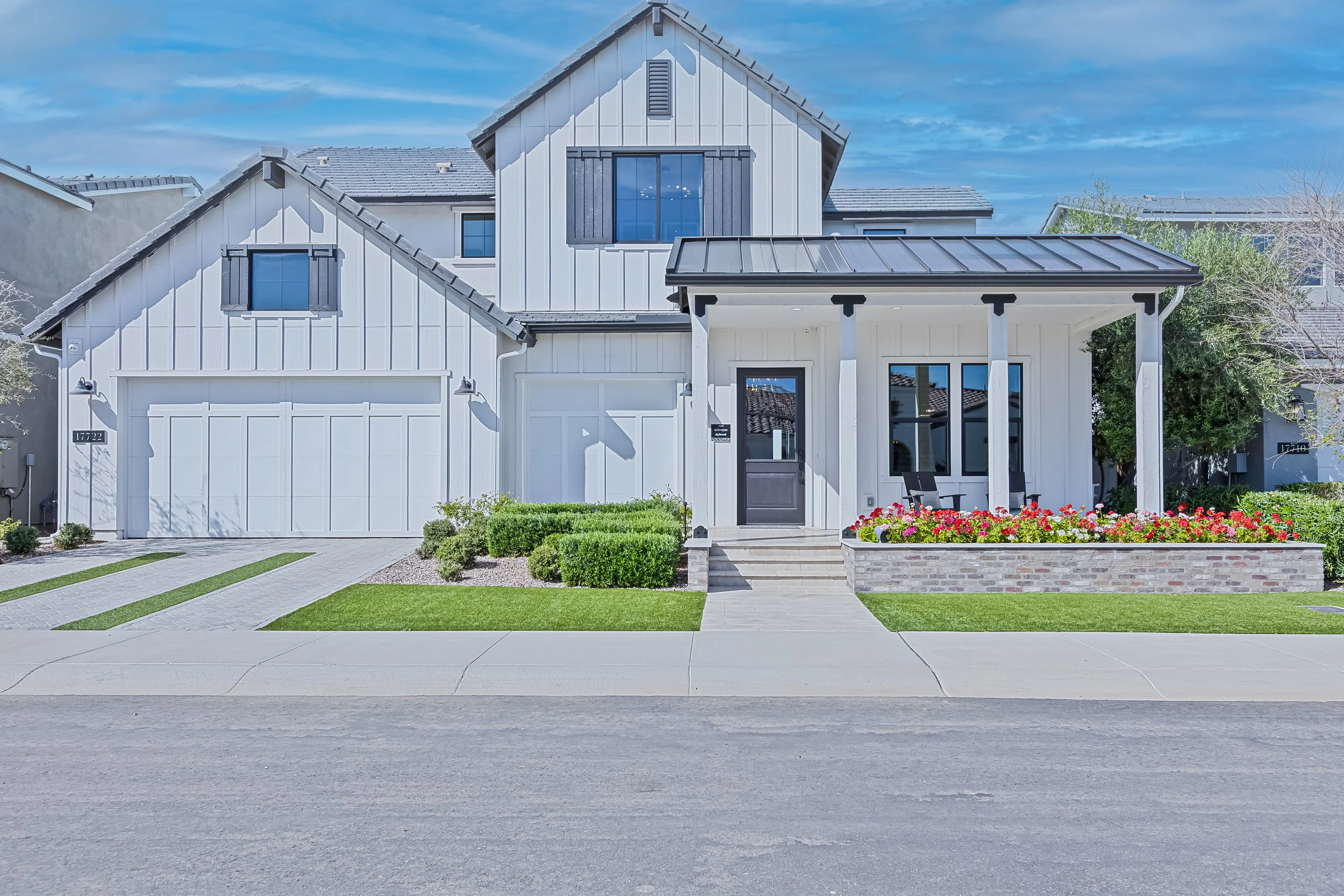 Modern white two-story house with a dark gray front door, black window shutters, and a landscaped front yard with grass, shrubs, and colorful flowers.
