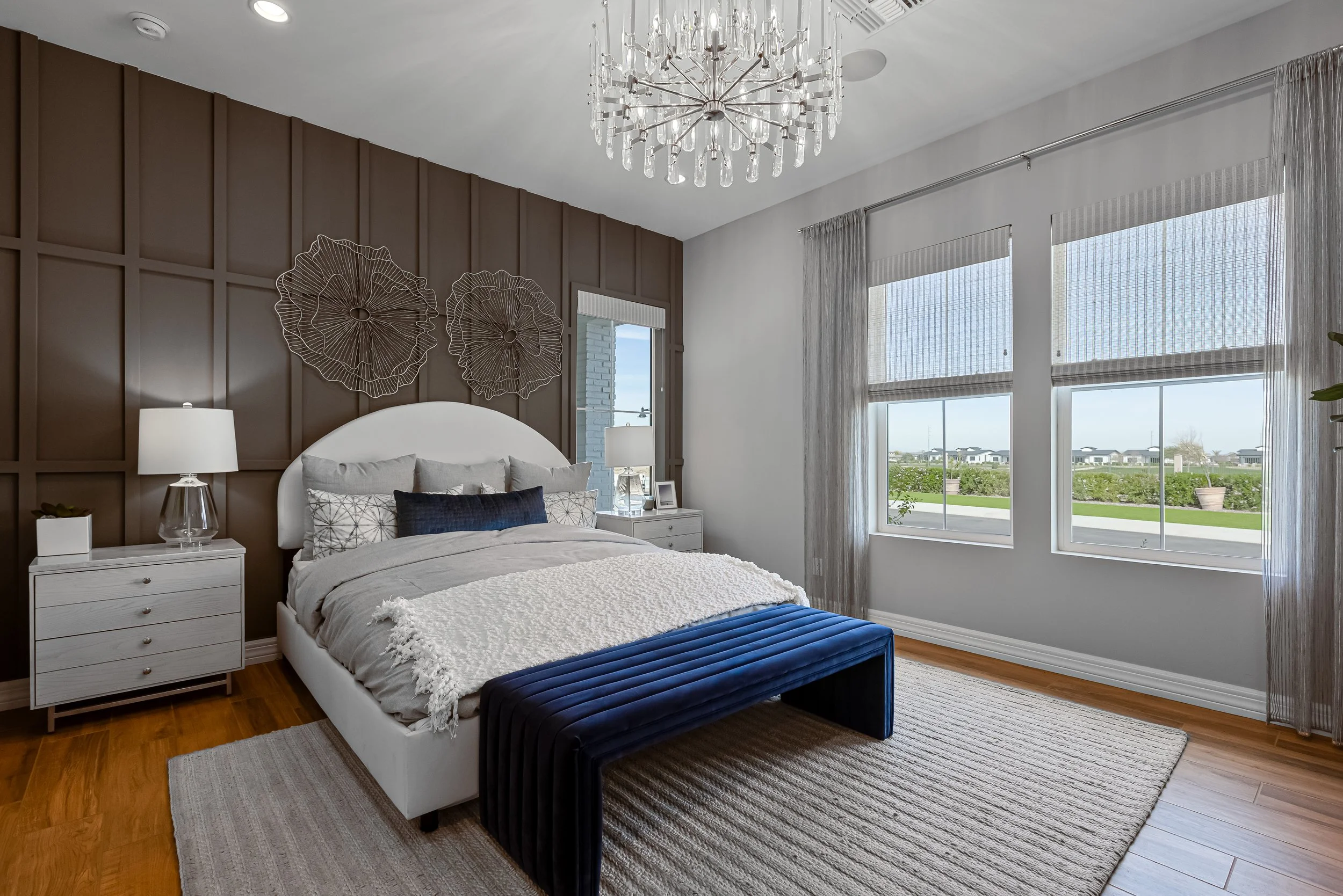 Modern bedroom with a large window, with a white bed featuring multiple pillows and a plush blue bench at the foot. Dark wood panel accent wall behind the bed, decorated with metal flower wall art. Two white nightstands with lamps and a small plant o