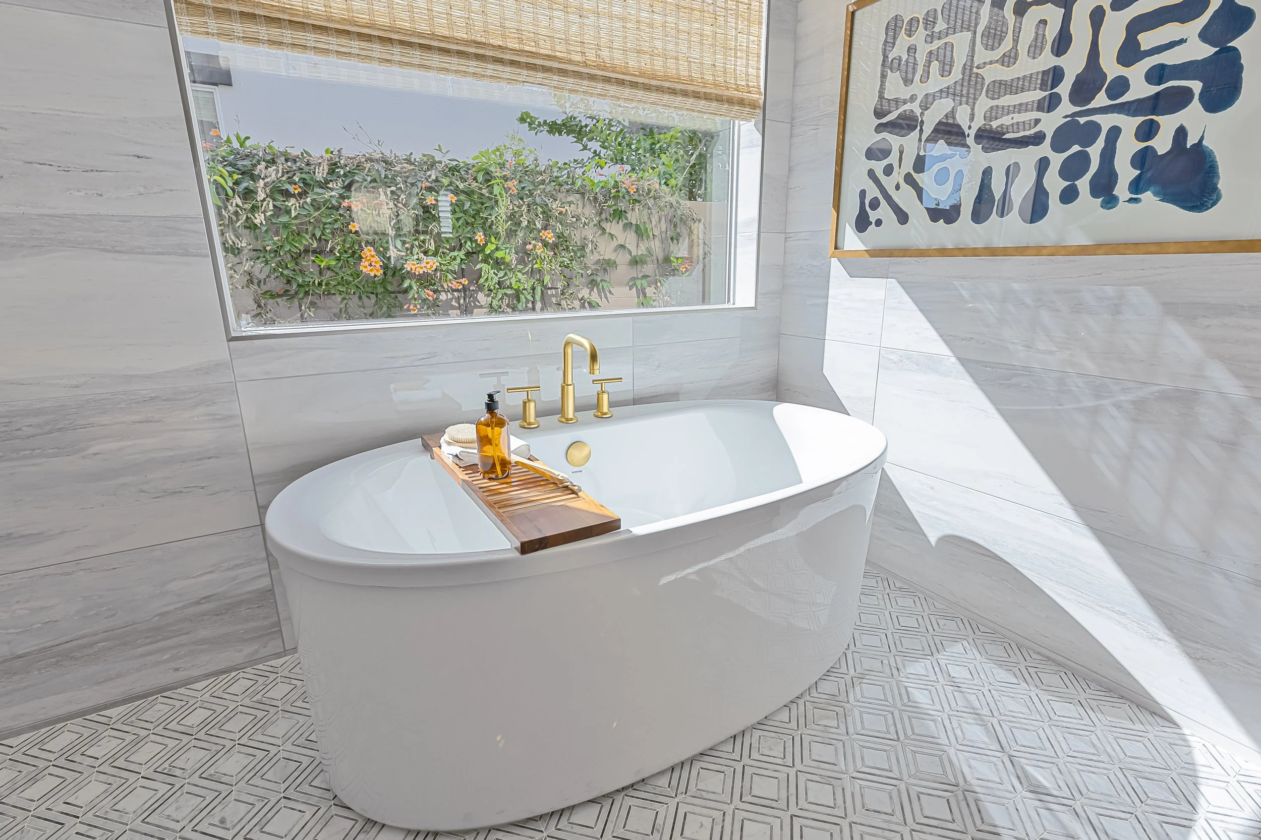 A modern white freestanding bathtub with gold fixtures in a bright bathroom, with a large window showing greenery outside, a wooden tray with soap and lotion, and abstract art on the wall.