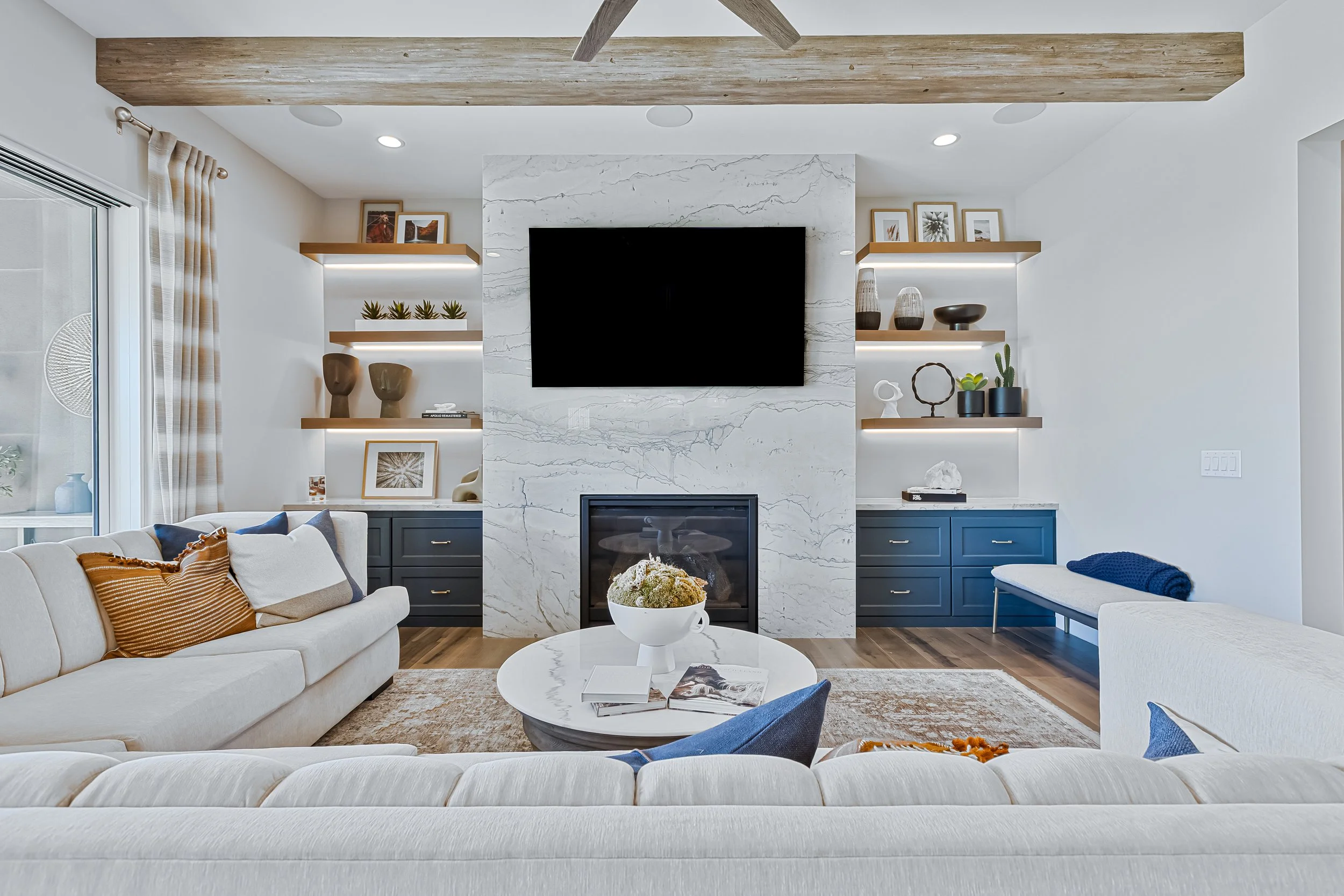 Modern living room with white sofas, a marble fireplace, a wall-mounted flat-screen TV, and built-in shelves with decor items and photos.