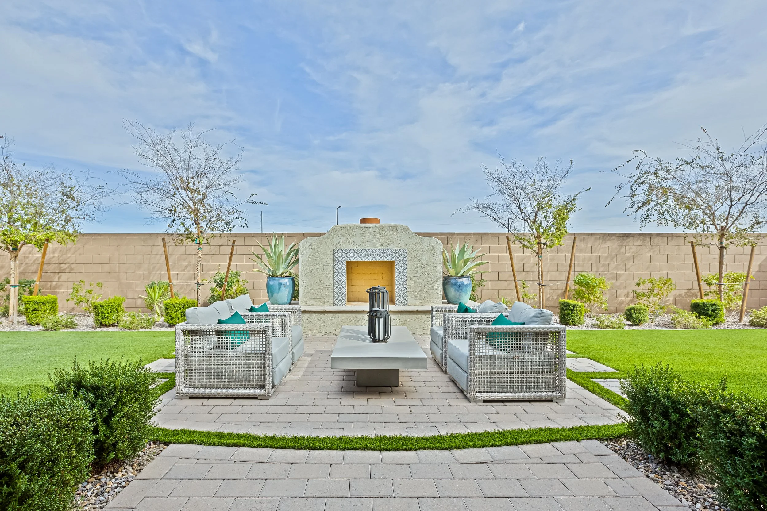 A modern outdoor patio with two white wicker sofas and teal cushions, a white coffee table with two black lanterns, and a fireplace with large potted plants on either side, set against a low fence and a landscaped garden with trees and bushes.