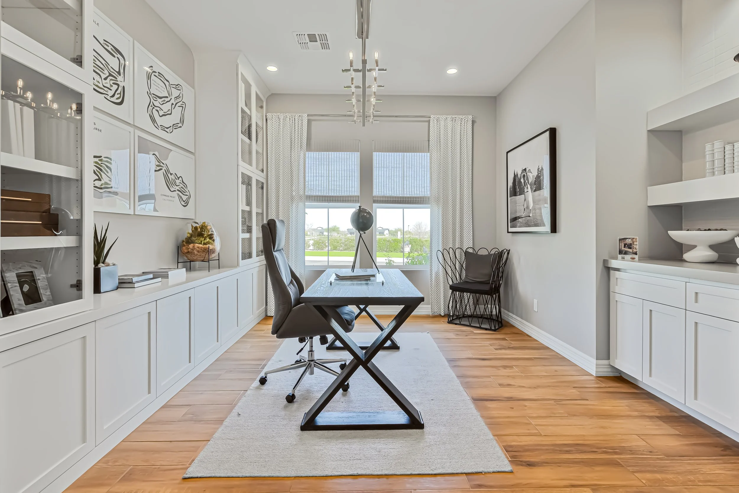 Bright home office with wooden floor, white cabinetry, and large window behind desk, decorated with plants, art, and modern lighting.