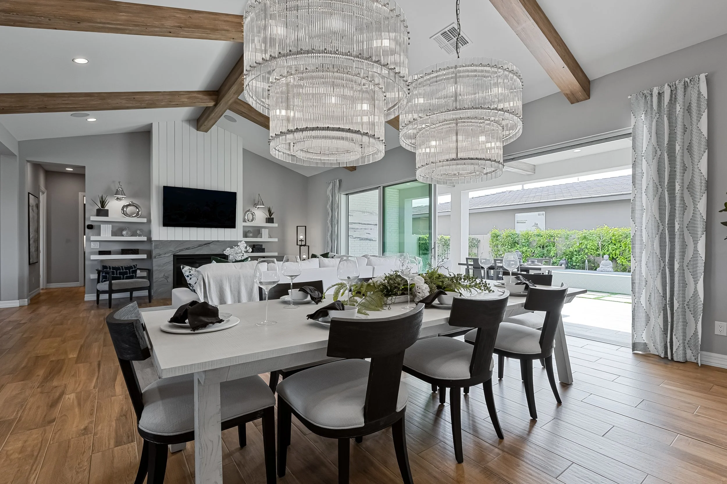 Modern dining room with a white table set with black napkins, wine glasses, and a floral centerpiece. Overhead, three large crystal chandeliers hang from a ceiling with wooden beams. Behind the table, there's a sitting area with a white couch and a f